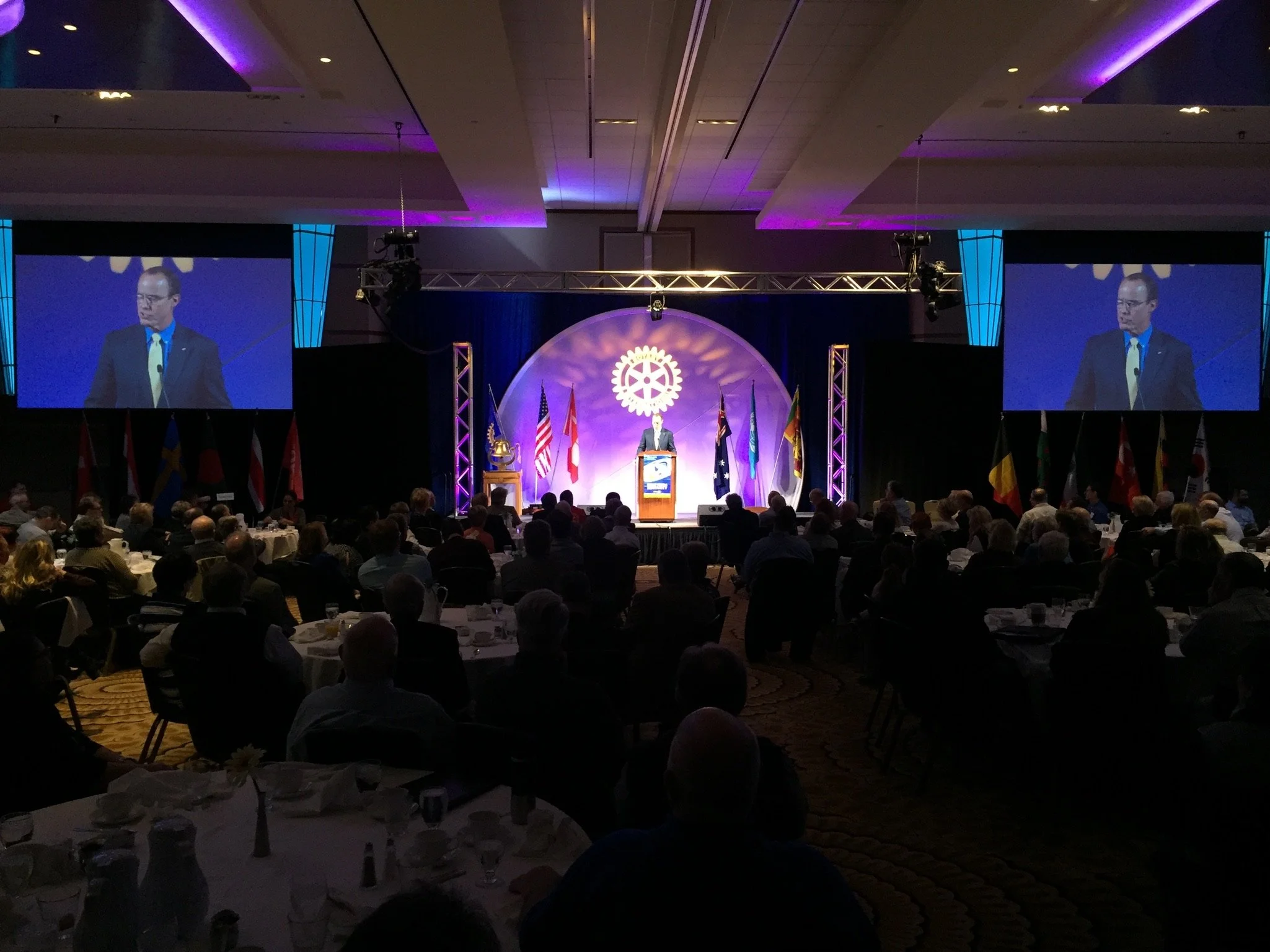 A man in a suit giving a speech at a conference stage with large screens on each side showing his image. The stage backdrop features a Rotary International emblem and flags. Audience seated at tables are visible in the foreground.
