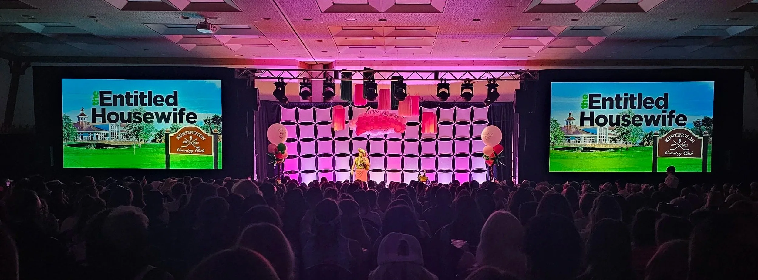 Large event set up with rear projection, stretch-shape backdrops, truss holding professional lighting to paint the room pink. Crowd awaiting event to start, event at Boyne Mountain Resort in Northern Michigan. Audio visual and magaged av.