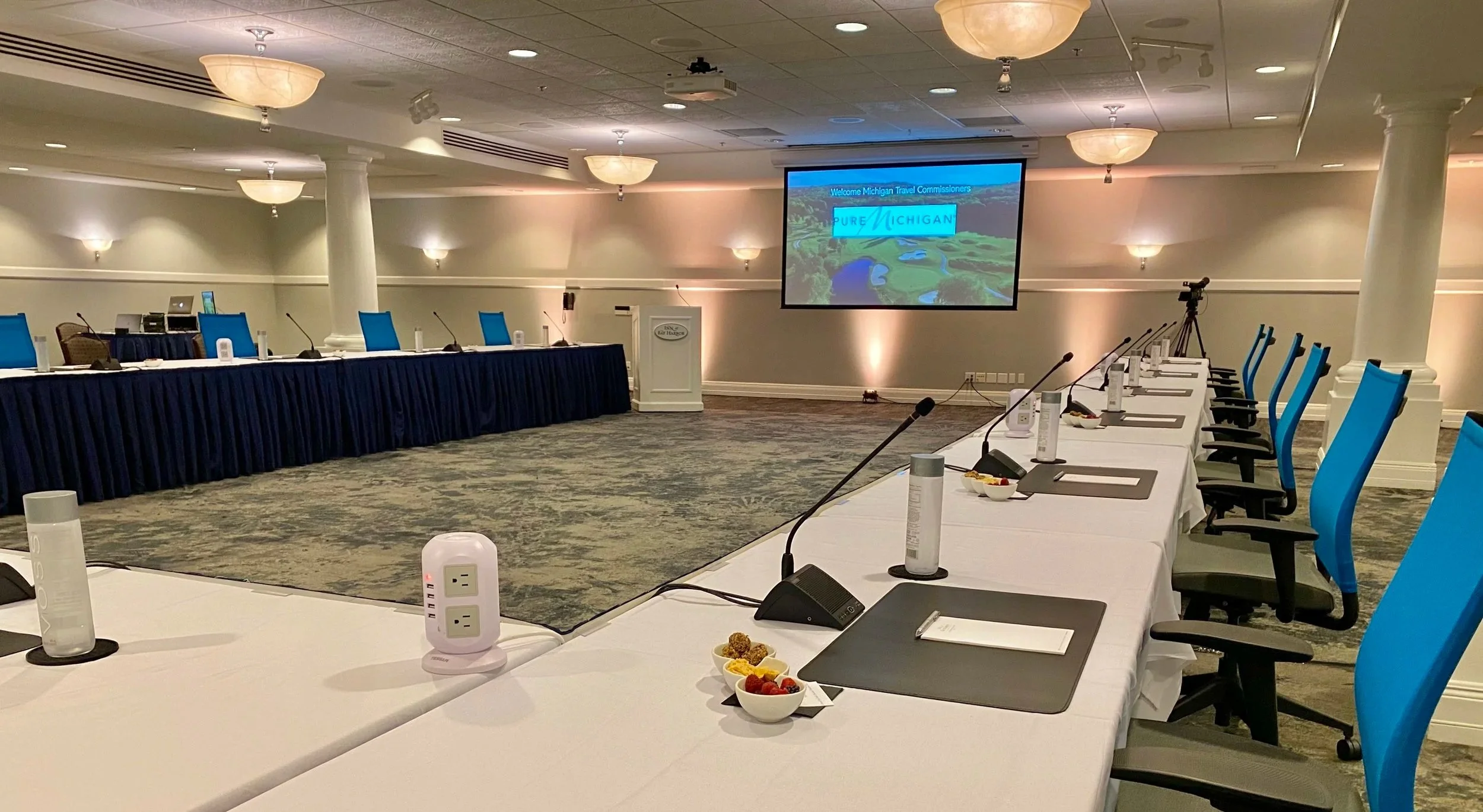 Conference room set up for a meeting with a U-shaped table arrangement, microphones, laptops, OWL. Installed large screen displays a Welcome message for Michigan Travel Commissioners with the Pure Michigan logo in the background.