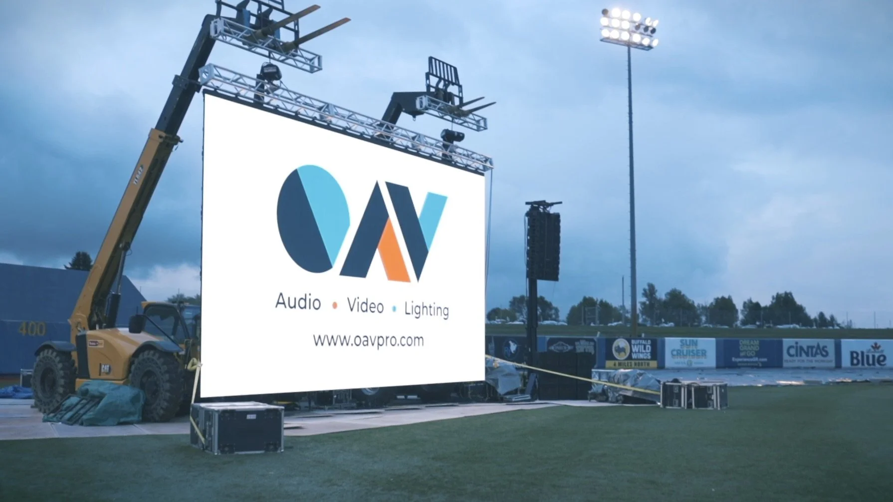 Large outdoor LED screen displaying a logo with the letters '', followed by the words 'Audio,' 'Video,' and 'Lighting,' and the website 'www.oavpro.com'. LED screen flown with cranes, large speaker and amps projecting sound to a sports stadium.