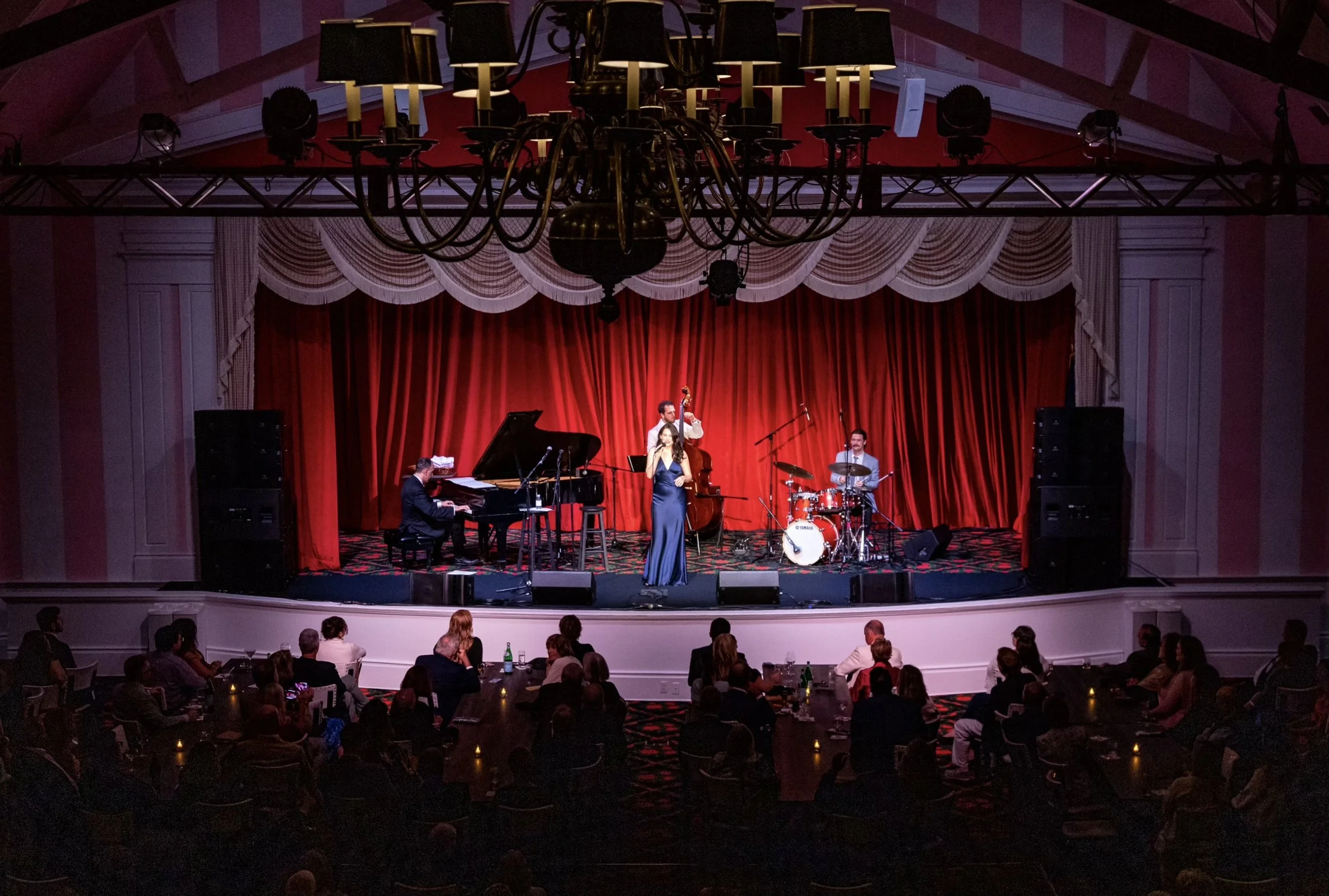 Live jazz band performing on Mackinac Island's Grand Hotel's Theatre stage with singer Stella Cole, pianist, double bassist, and drummer in front of an audience in a theater with red velvet curtains. 
