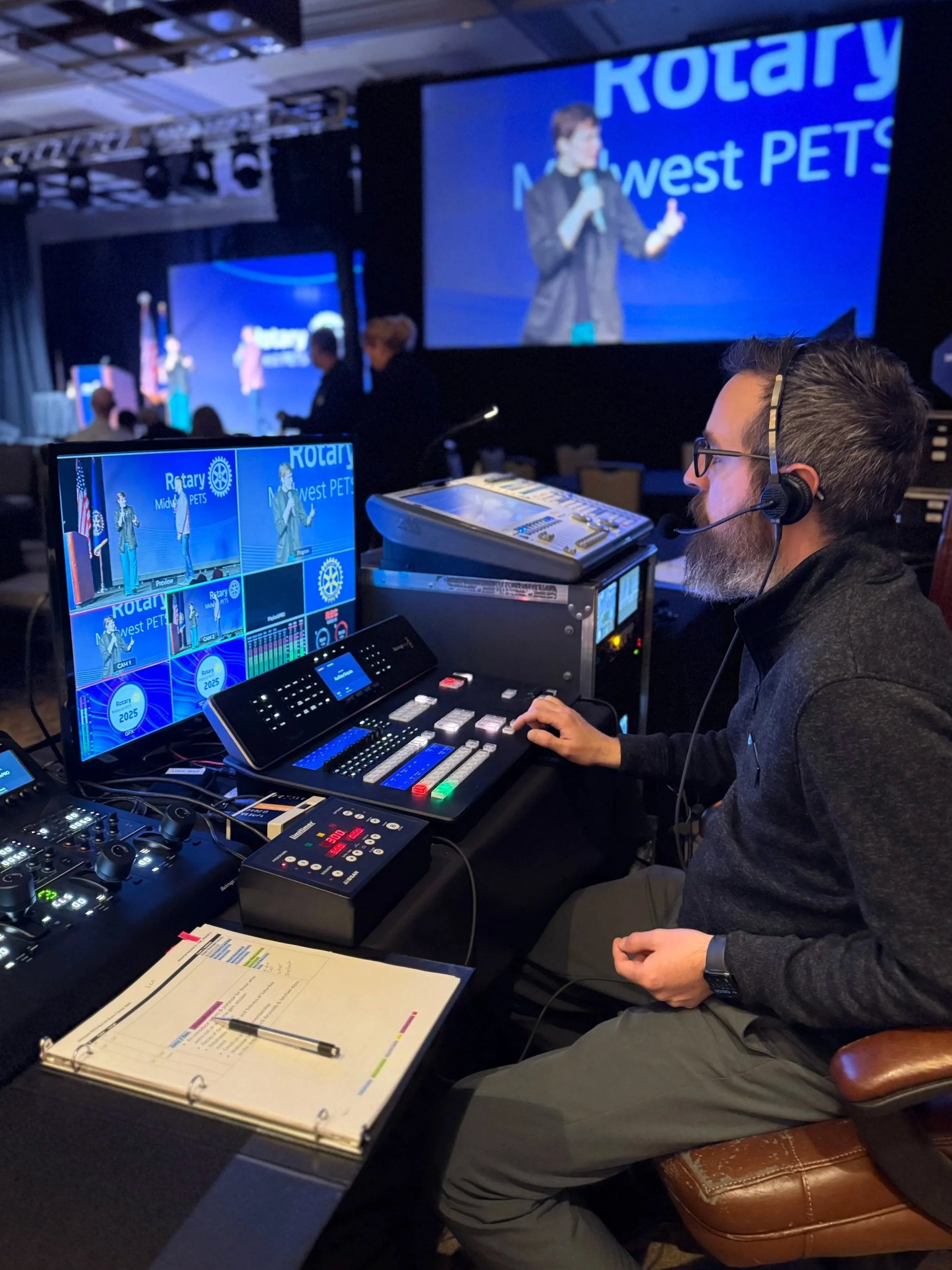Video director AV techncian wearing a headset working at a monitor at the AV station to the side of the general session of a large event with multiple projectors, LED video wall backdrop, lighting, staging, truss equipment for Rotary corporate event