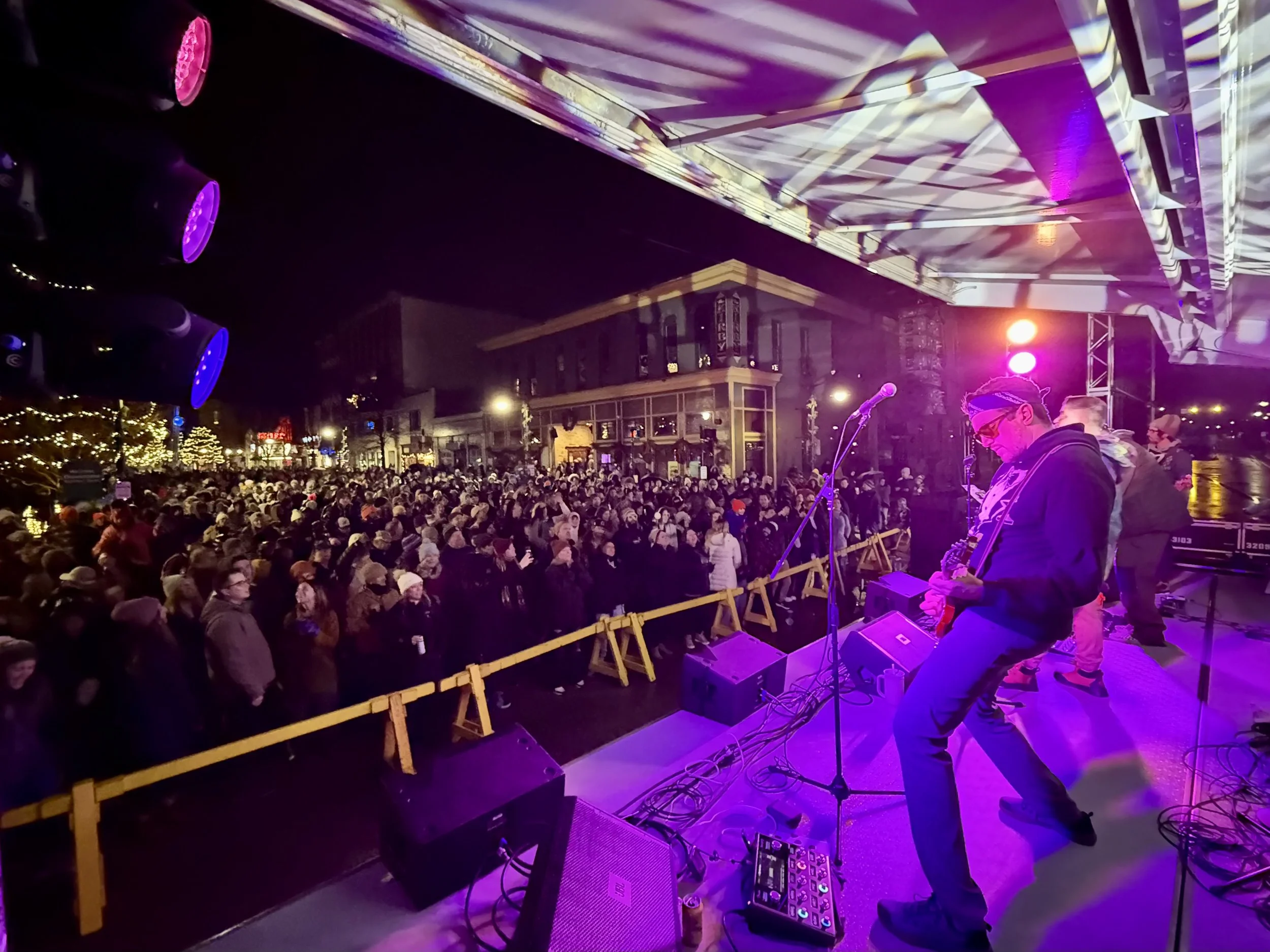A large crowd gathered at an outdoor concert at night. A musician is performing on stage with purple and pink wash lighting. Speakers, amplifiers, stage, LED video wall display to crowd. New Years Eve Concert in Grand Haven, West Michigan.