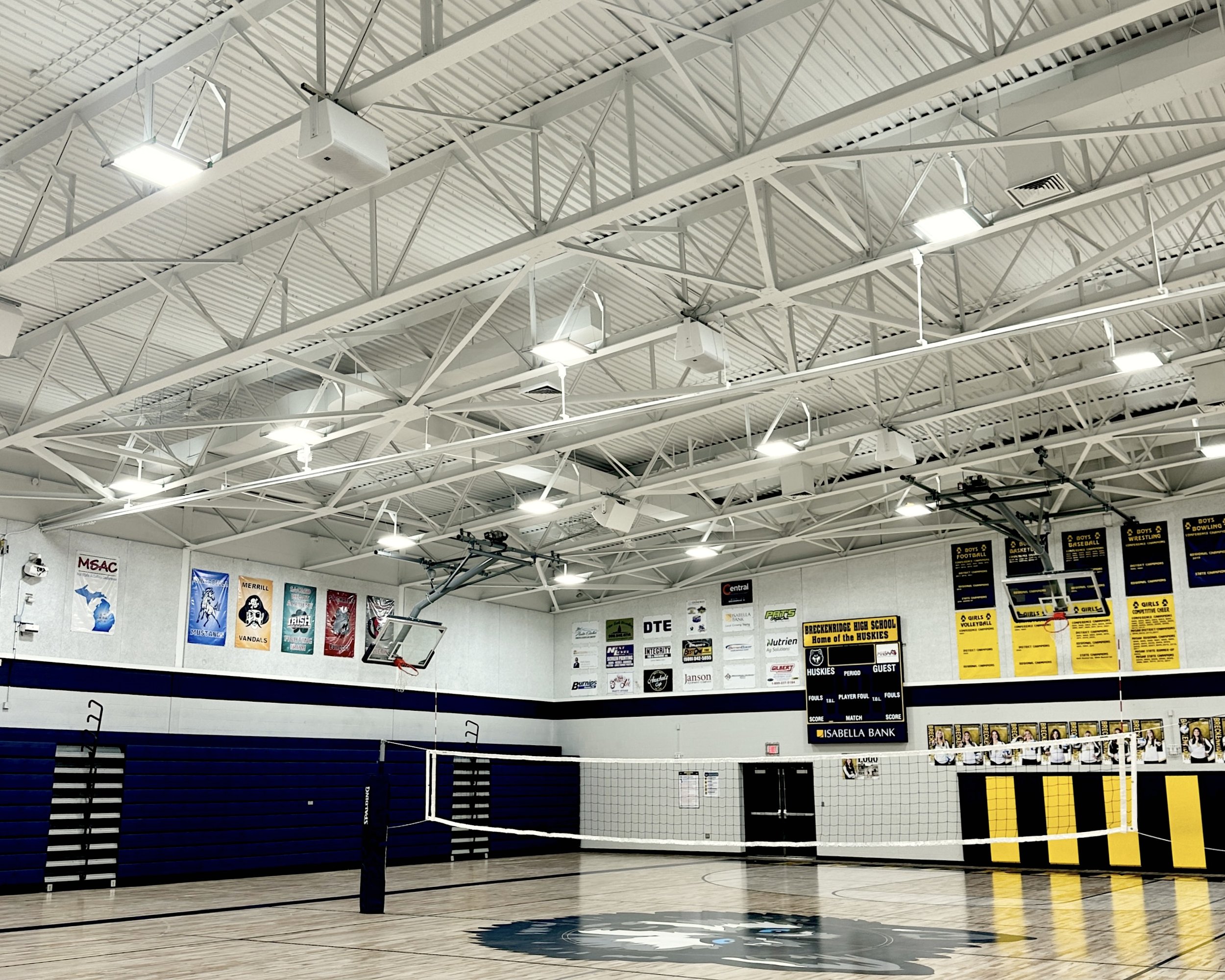 Breckenridge High School indoor basketball court inside a high school gymnasium with banners hanging on the walls and a scoreboard. Audiovisual installation of speaker and sounds, AV control console on the wall. Speakers blend in with ceiling.