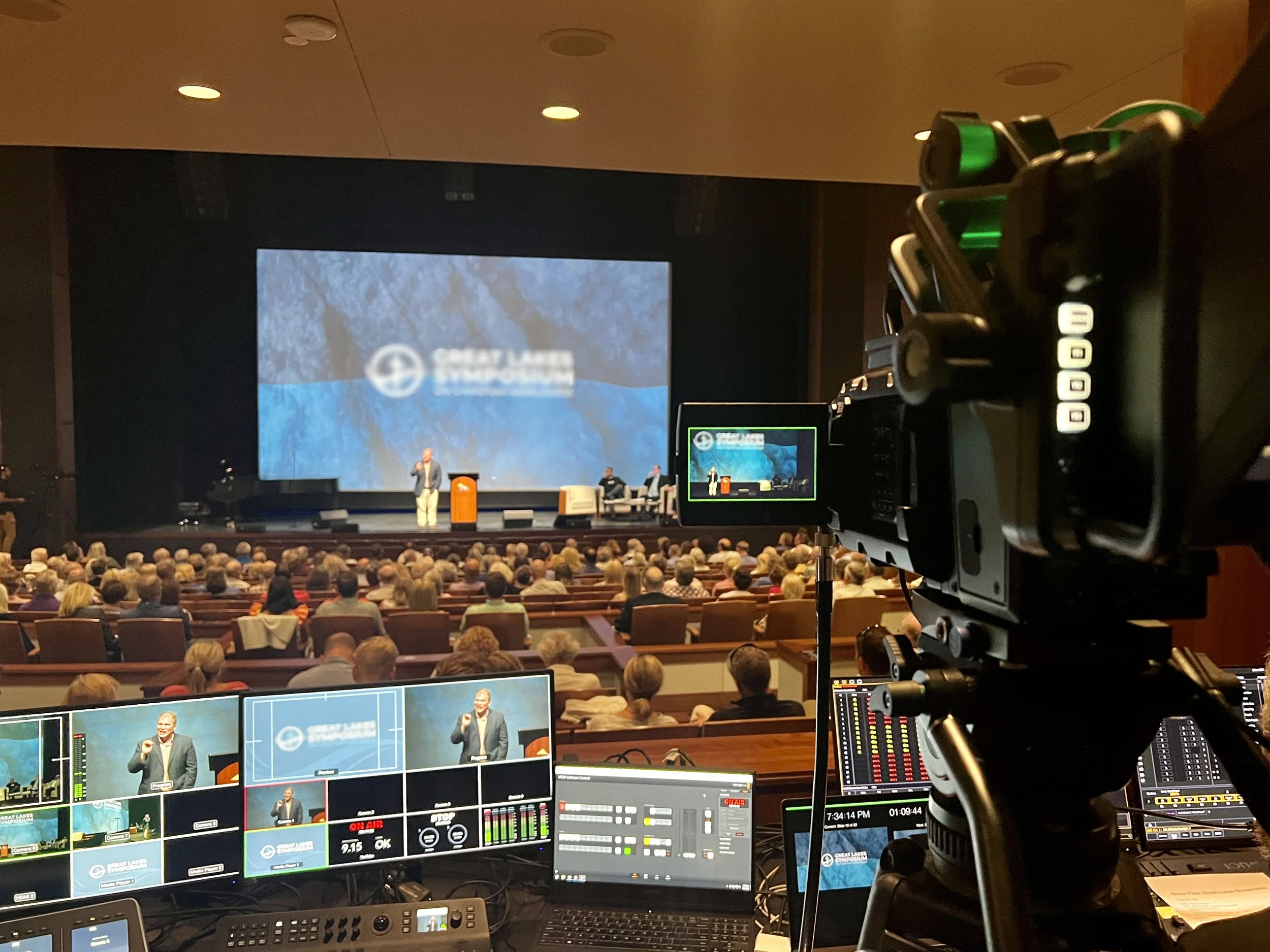 A stage with a speaker giving a presentation for Great Lakes Christian Symposium at Great Lakes Center for Arts in Bay Harbor Michigan, audience seated in front, with video, recording, and sound equipment in the foreground by audio visual company OAV