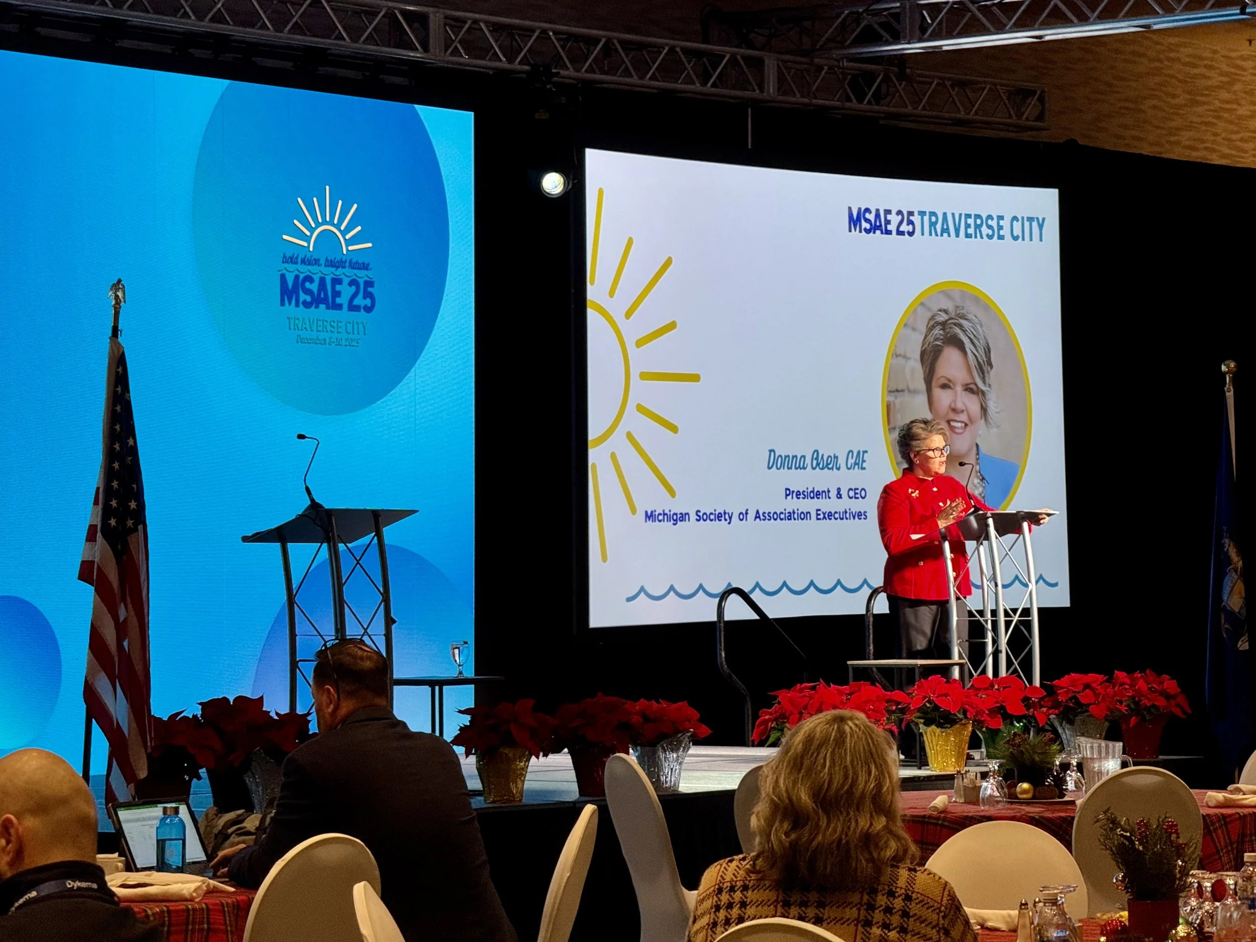 A woman standing at a lectern giving a speech at a conference. Behind her are screens and an LED wall with logos and presentation content. The Michigan Society of Association Executives' 25th anniversary celebration in Traverse City, December 2025.