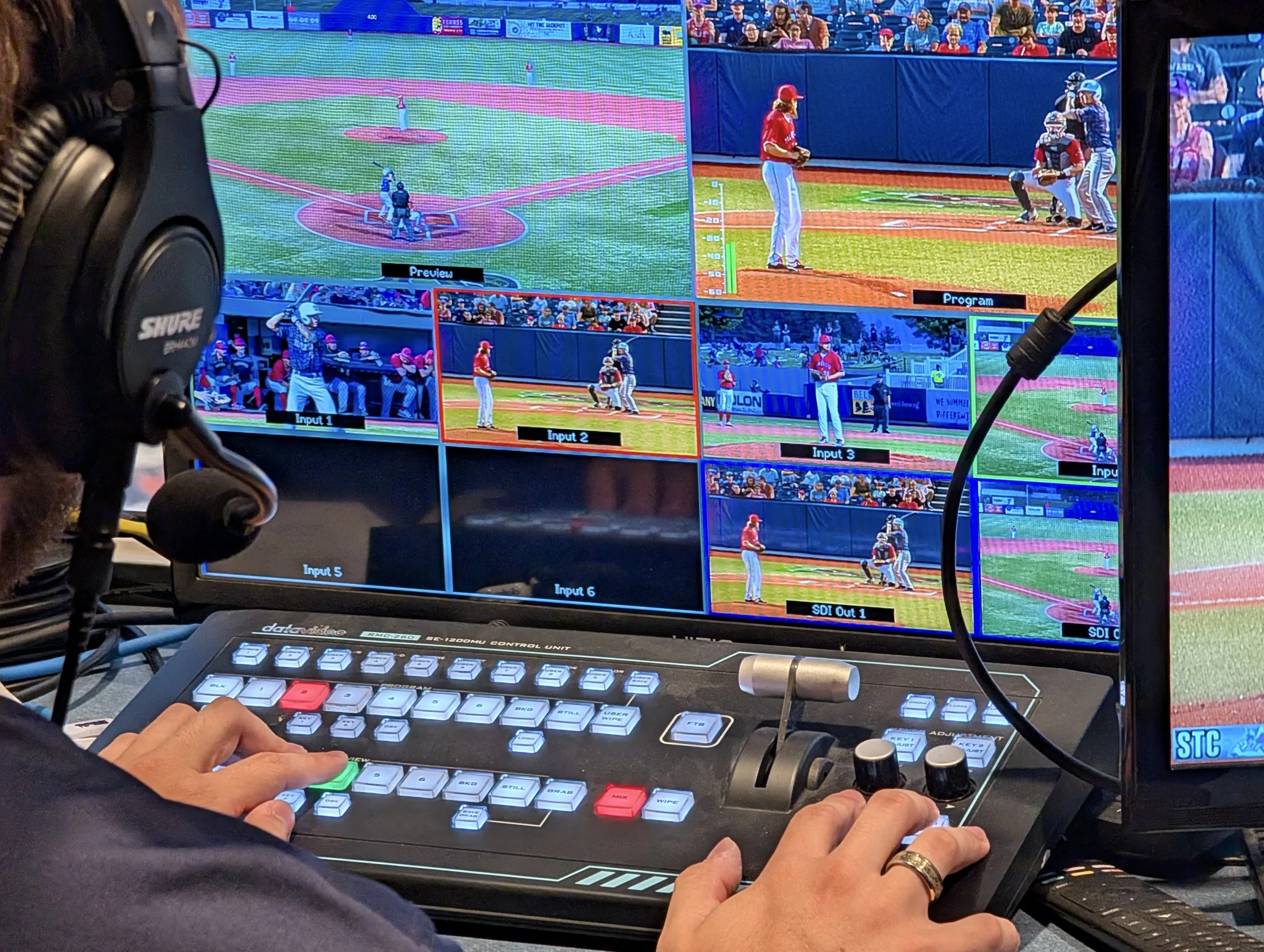 A video control room with a monitor displaying a live baseball game, focusing on the pitcher, batter, and catcher on the field. There are additional smaller screens below showing different camera angles, and a person operating a control panel with va