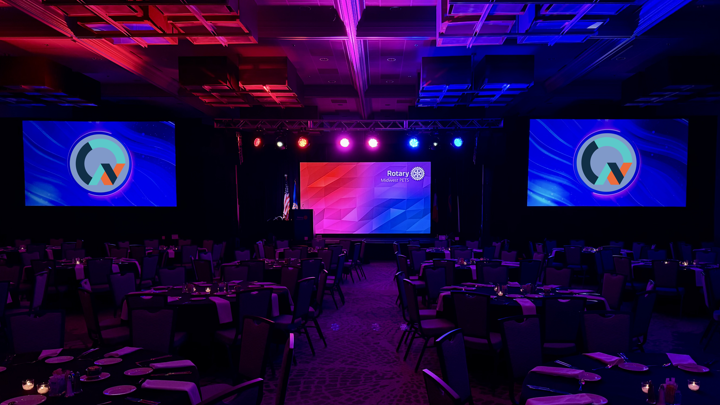 A large event ballroom with round tables set for dining, stage with two large screens displaying a logo, and colorful stage lighting in purple, blue, and pink. Martin pro lighting on tyler truss, general session to a national event.