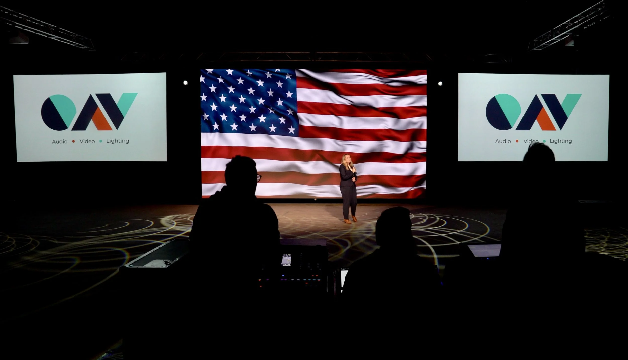 A woman speaker standing on a stage with an American flag backdrop on LED video wall, supported by two large front projection screens displaying a logo with text: Audio, Video, Lighting. AV Technicians manning av table and entire setup.