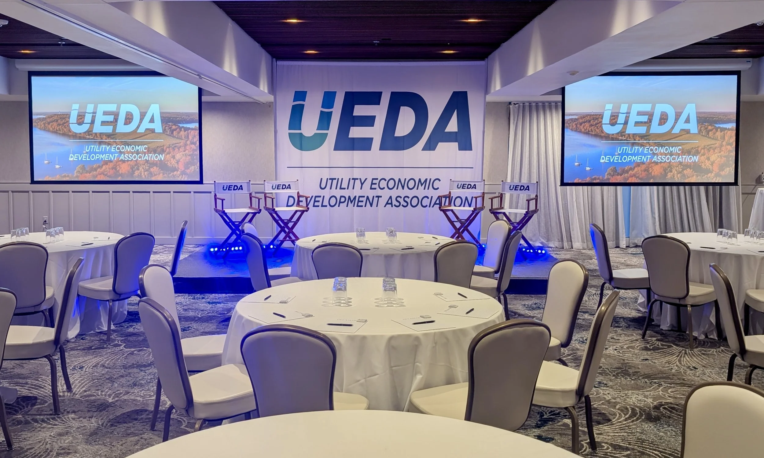 Conference room setup with round tables and chairs, a stage with dual large screens displaying the UEDA logo, and banners with the organization's name and slogan. Managed AV provided for Delamar Hotel.