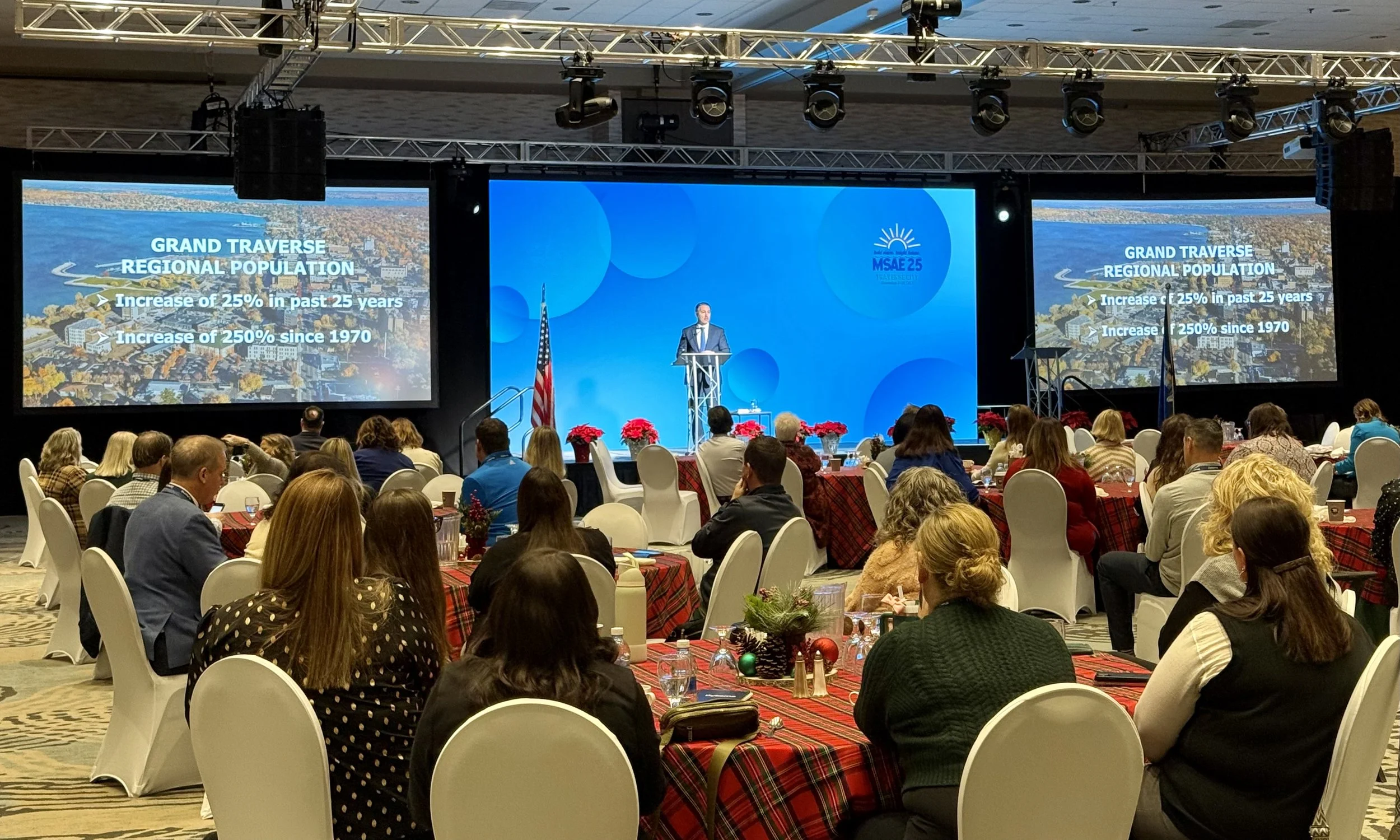 A conference room filled with people sitting at round tables with red and black plaid tablecloths. There are small Christmas decorations on the tables. A speaker in a suit stands at a podium on a stage, delivering a presentation. Two large screens on