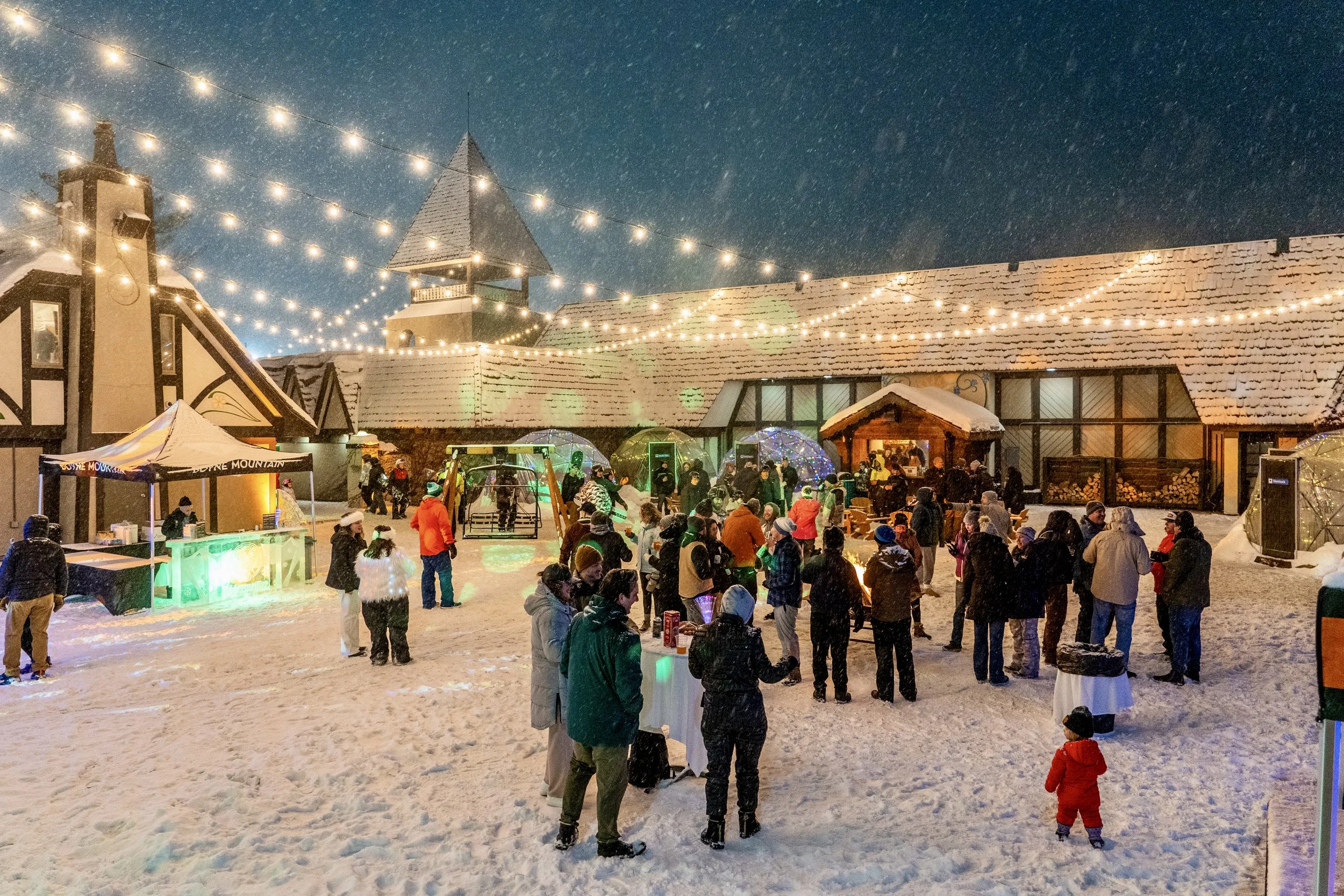 A festive winter scene at night with string lights hanging above a snow-covered outdoor area filled with people. Ice Apres at Boyne Mountain Resort 2025, Blizzard Uplighting in ice displays, wash lighting projected down on crowd and space.