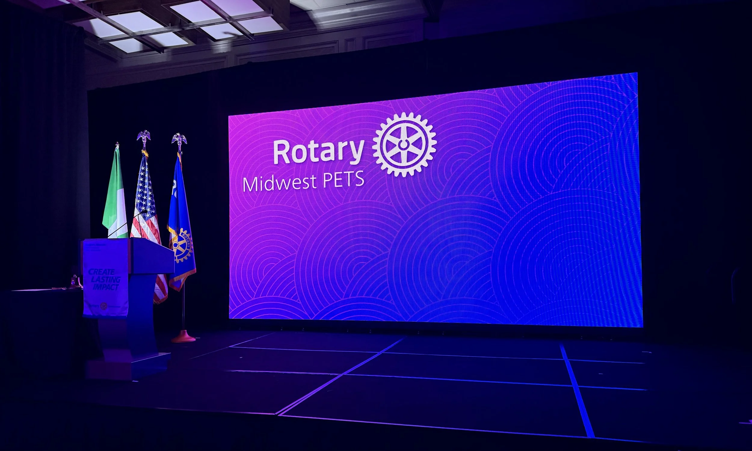 Rotary event breakout session stage with a large LED video wall displaying the Rotary Interlogo and the text 'Rotary Midwest PETS' in purple, with American, Italian, and Rotary flags on the left side, and a podium with a Rotary banner in front of it.