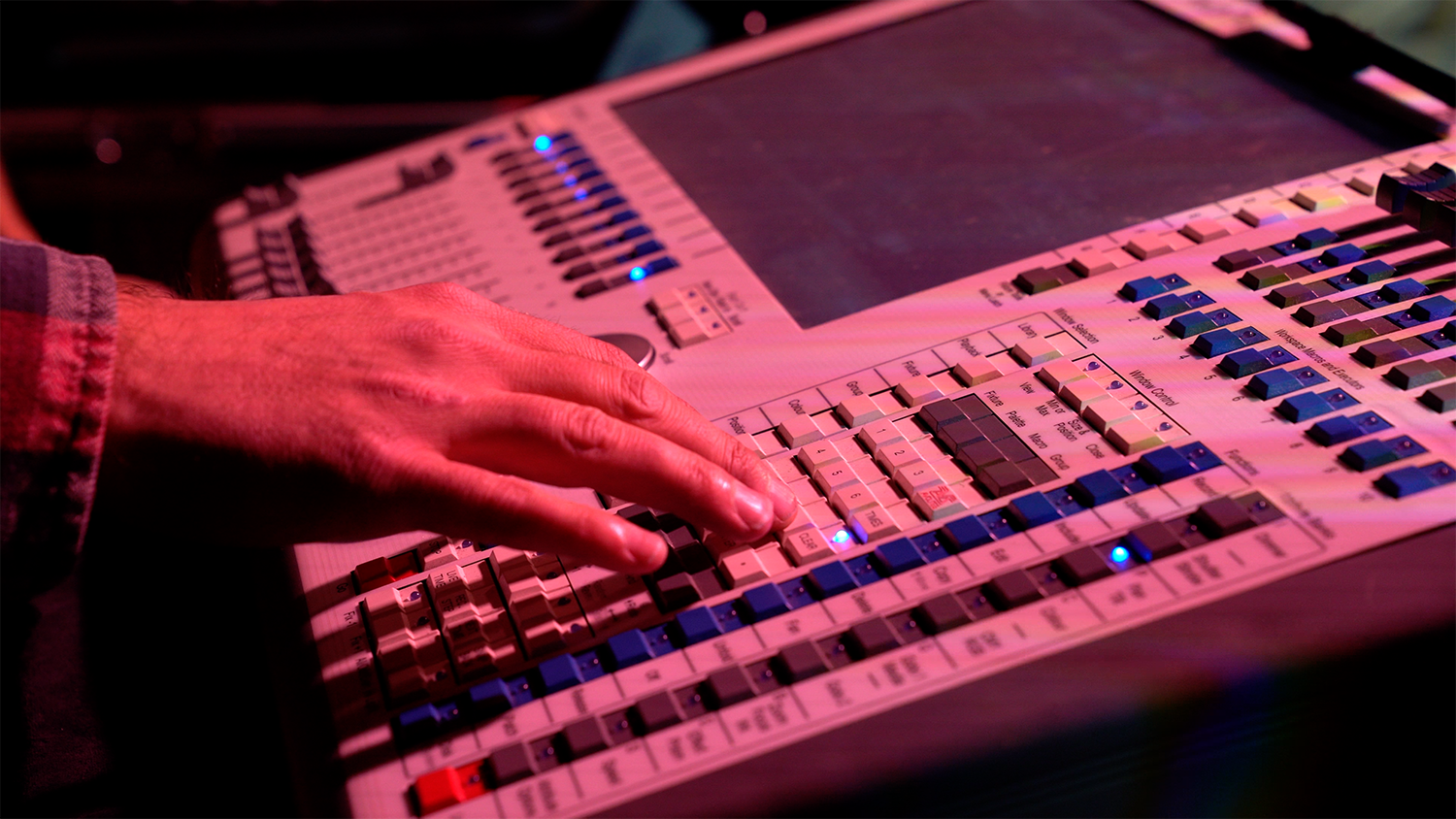 Professional audiovisual technician operating a professional sound mixing console avolite tiger touch with numerous sliders, knobs, and a digital display, lit by pink and purple LED stage lights.