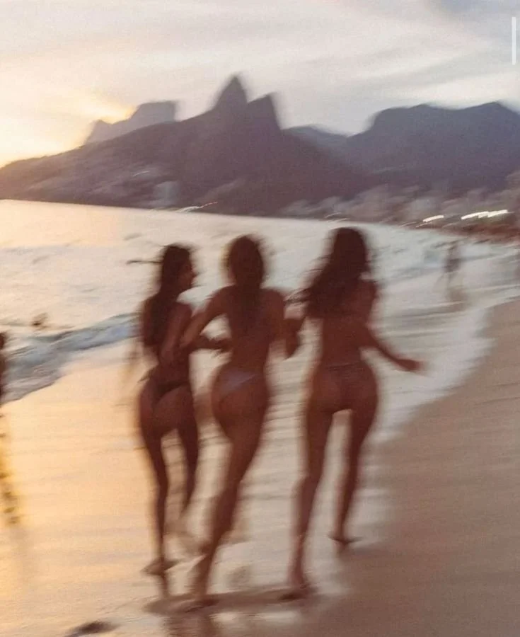 Three women in swimsuits walking along the beach at sunset with mountains and water in the background.