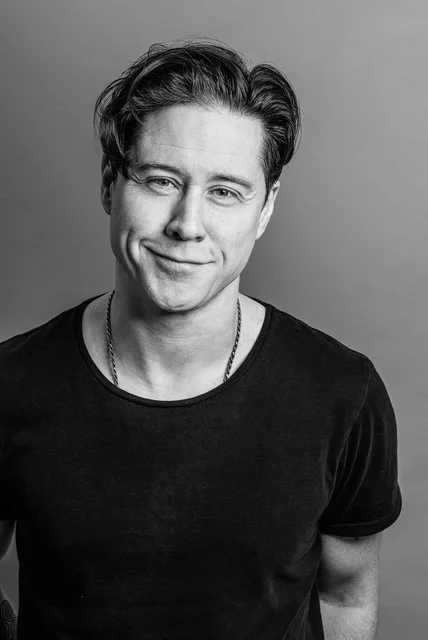 A young man with dark hair, smiling with a slight tilt of the head, wearing a black T-shirt and a chain necklace, in front of a plain background.