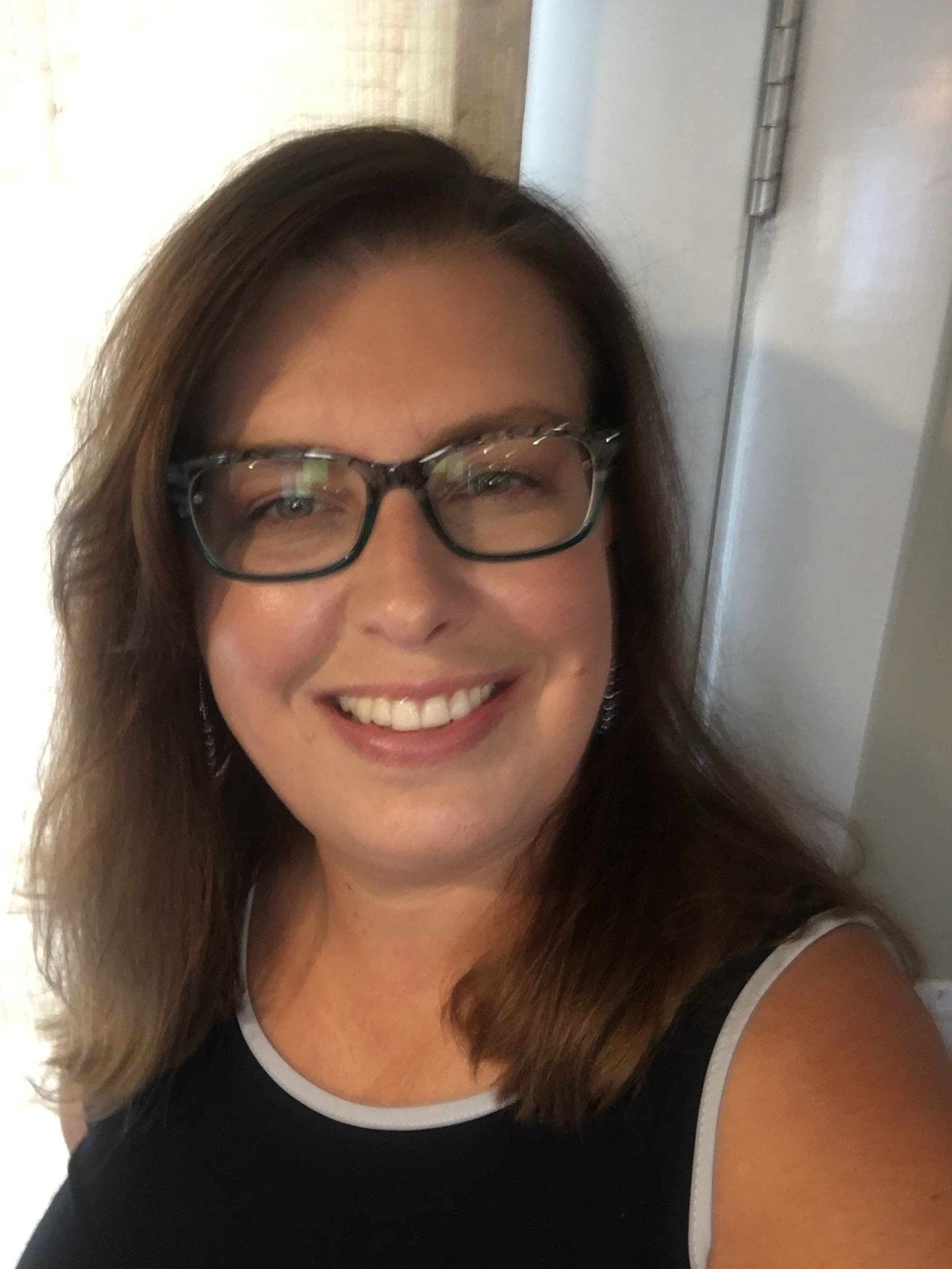 A woman with glasses and shoulder-length brown hair smiling indoors.