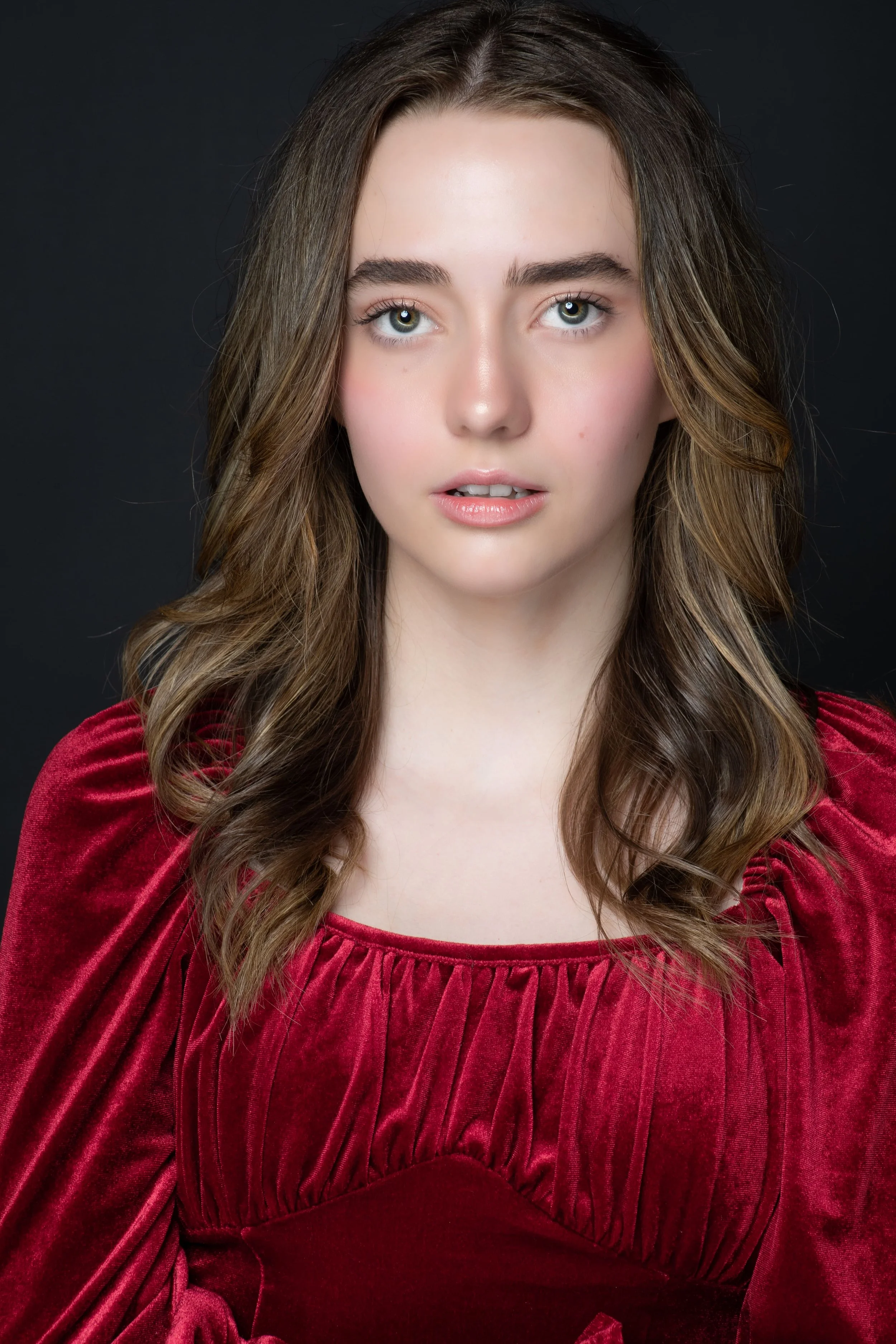 A young woman with long wavy brown hair, wearing a red velvet top, looking directly at the camera against a dark background.