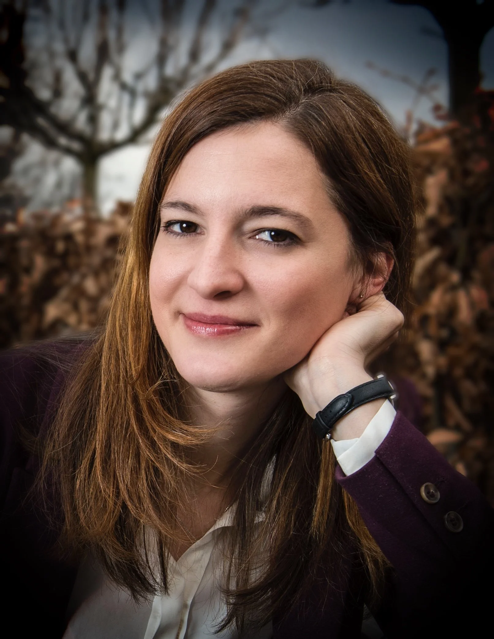 A woman with brown hair and fair skin, wearing a white shirt and dark blazer, resting her head on her hand outdoors with trees and a cloudy sky in the background.