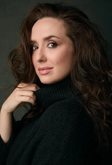 A woman with long, wavy brown hair and light skin, wearing a black turtleneck sweater, looking at the camera with a slight smile.