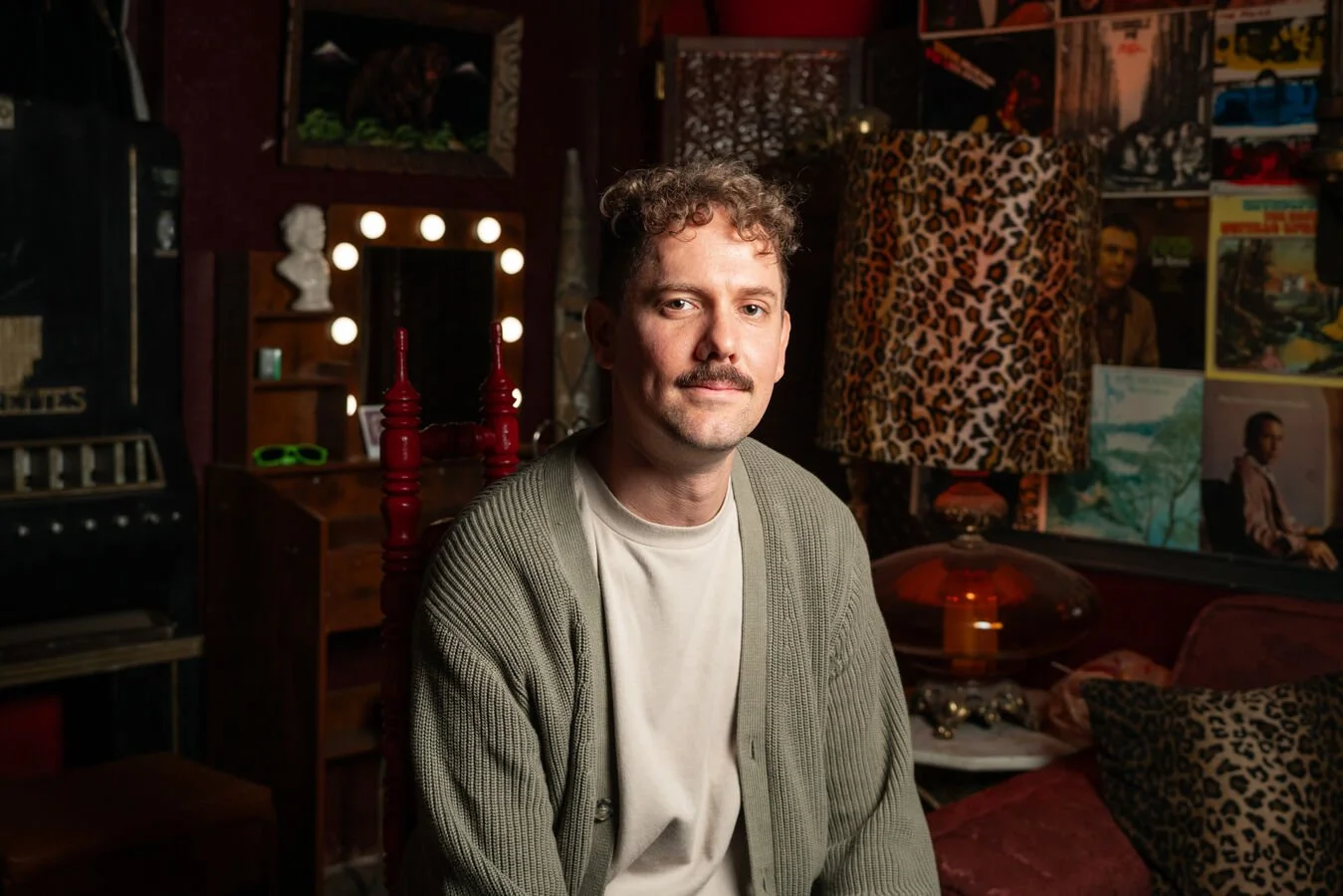 A young man with curly hair and a mustache sits in a dimly lit, eclectic room filled with vintage decor, paintings, and a leopard print lampshade.