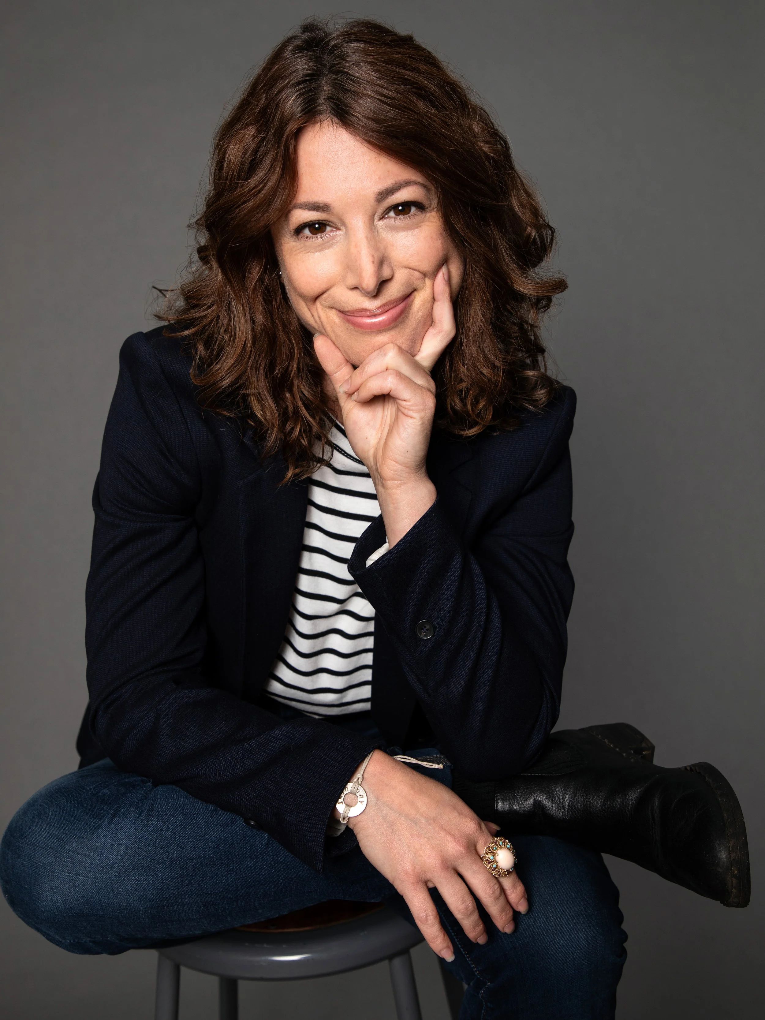 A woman with wavy brown hair, wearing a navy blazer, striped shirt, jeans, and black boots, sitting on a stool and smiling thoughtfully at the camera.