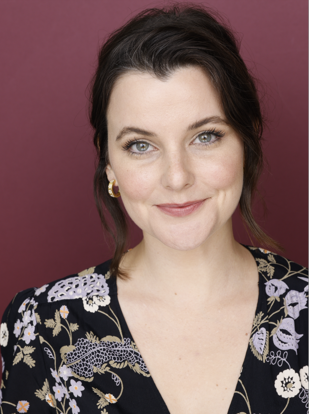 A woman with shoulder-length dark hair, green eyes, and fair skin smiling against a maroon background. She is wearing a black top with floral embroidery and gold hoop earrings.