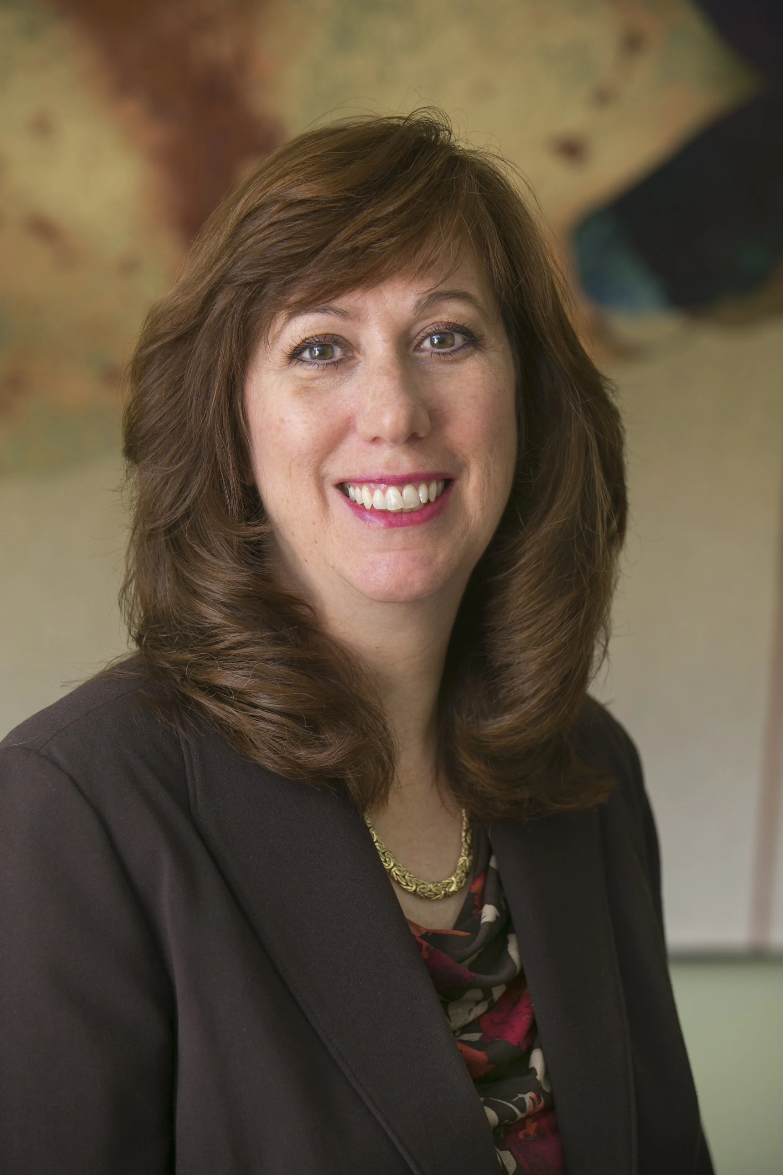 A woman with medium-length brown hair, wearing a black blazer and a patterned top underneath, smiling with visible teeth, and a gold necklace. The background appears to be an abstract painting.