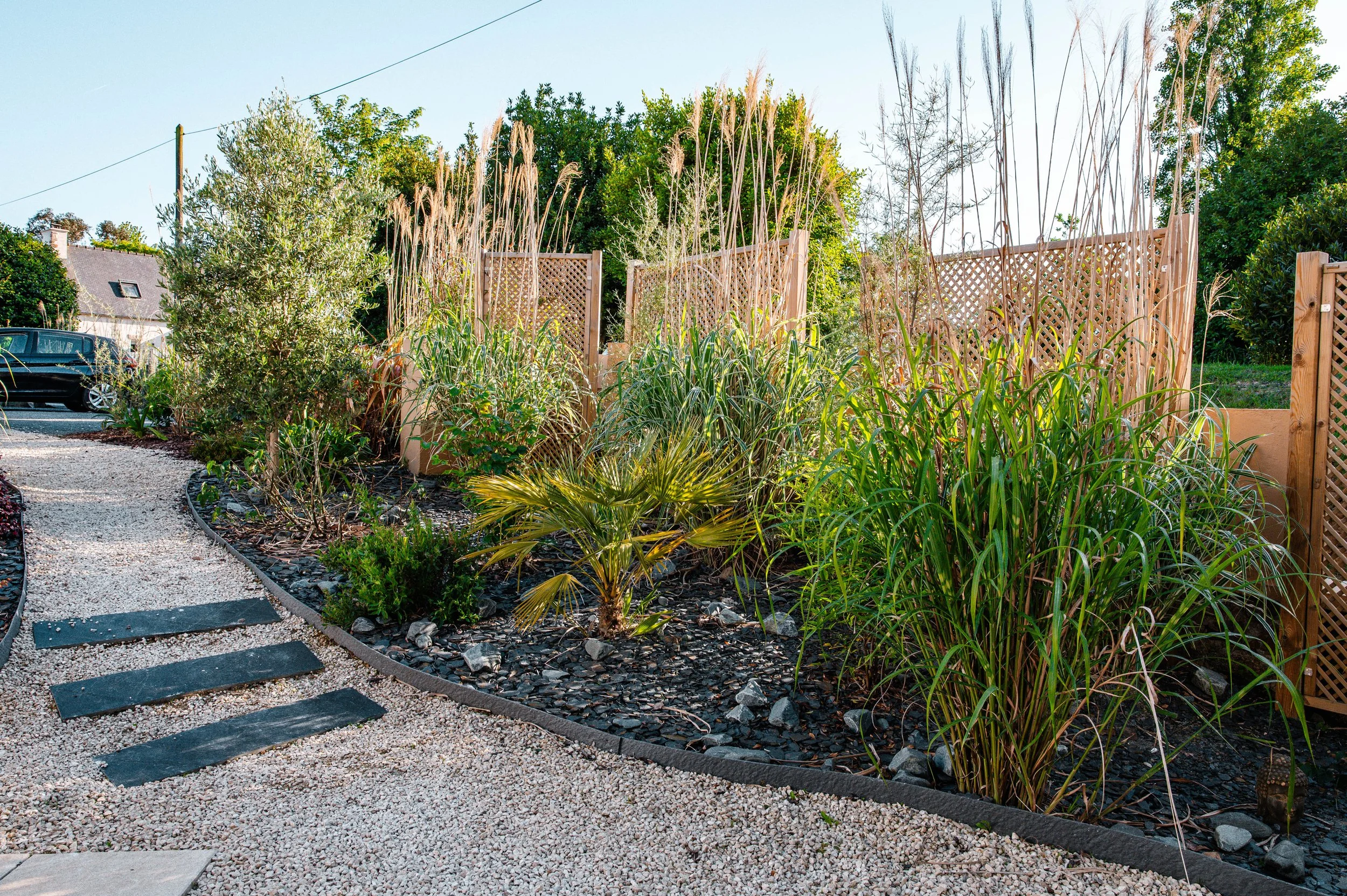 Jardin paysager avec un sentier en gravier, plantations de buissons et d'herbes hautes, clôture en bois décorative, et un arbre à gauche, sous un ciel clair.