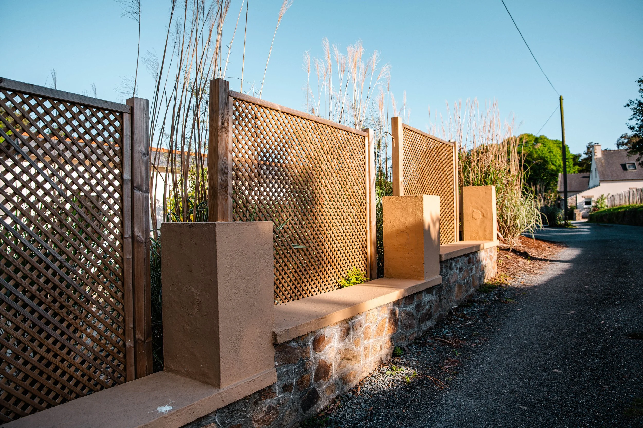 Clôture en bois avec pierre de base, bordure de chemin asphaltée, arbres et maisons en arrière-plan, ciel bleu.
