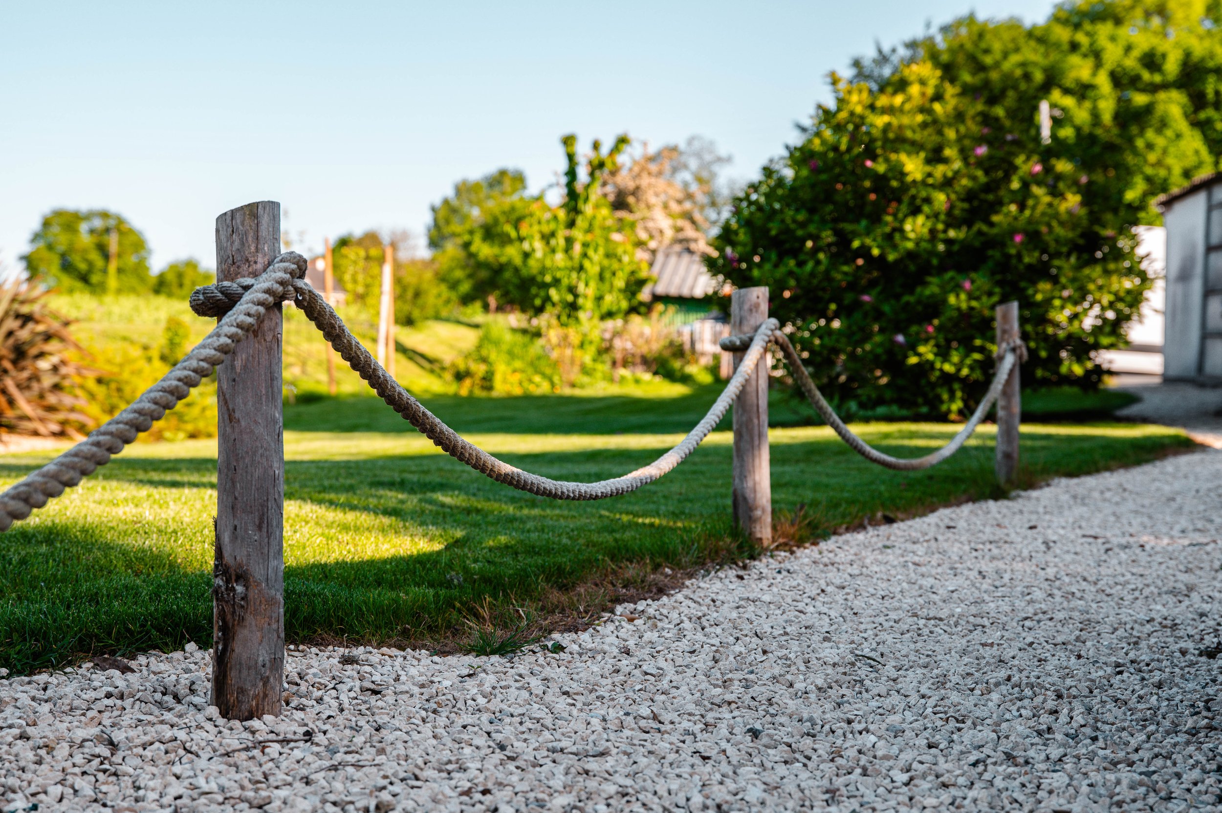 Clôture en corde avec des poteaux en bois séparant une pelouse verte et un chemin en gravier, avec des arbres et des bâtiments en arrière-plan en plein jour.