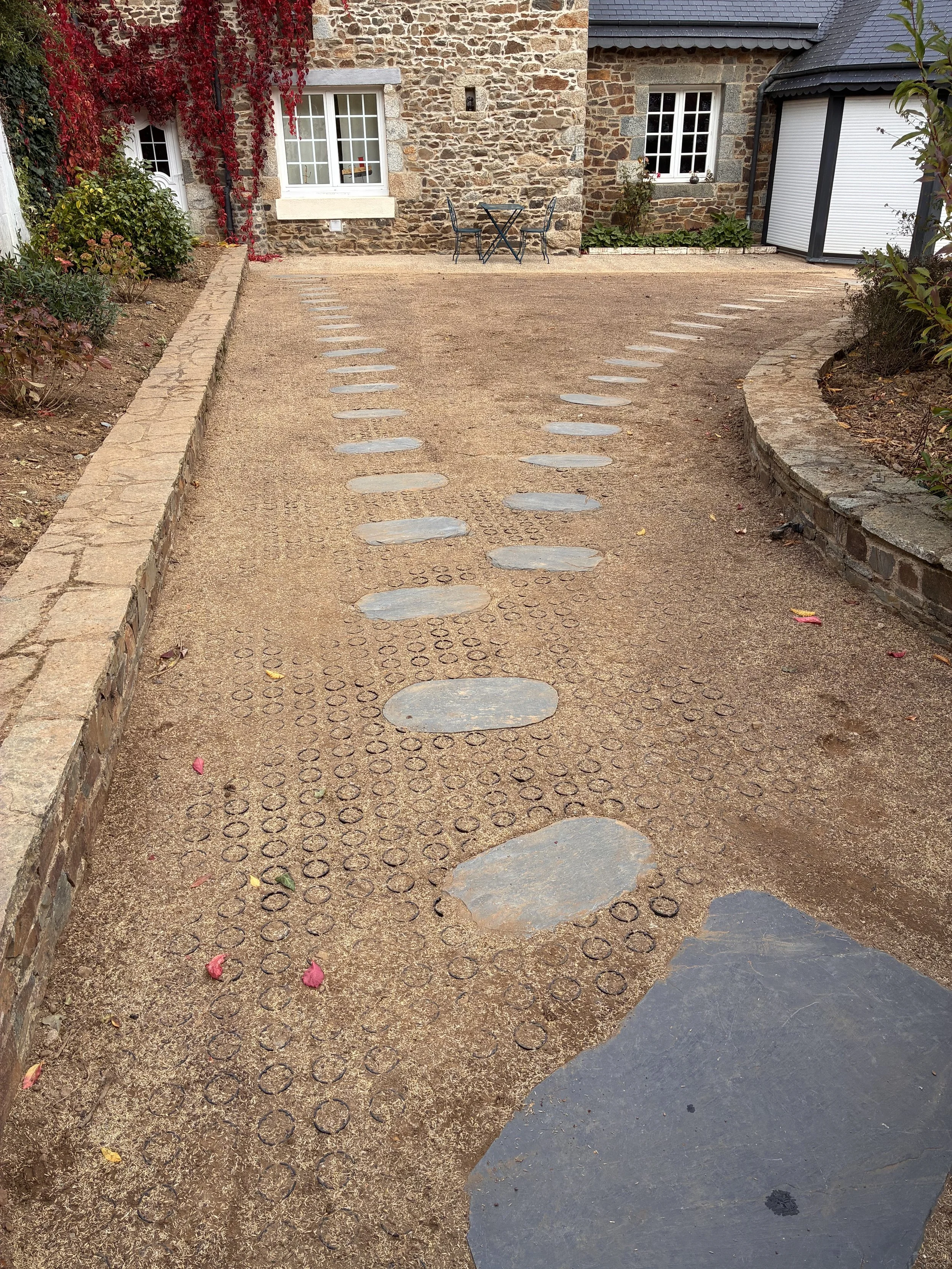 Allée en gravier menant à une maison en pierre avec des fenêtres à carreaux blancs, entourée de plantes et de fleurs, avec des pierres plates incrustées dans le sol.