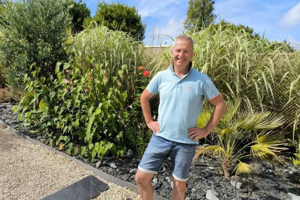 Homme souriant debout dans un jardin avec des plantes vertes, sous un ciel bleu.