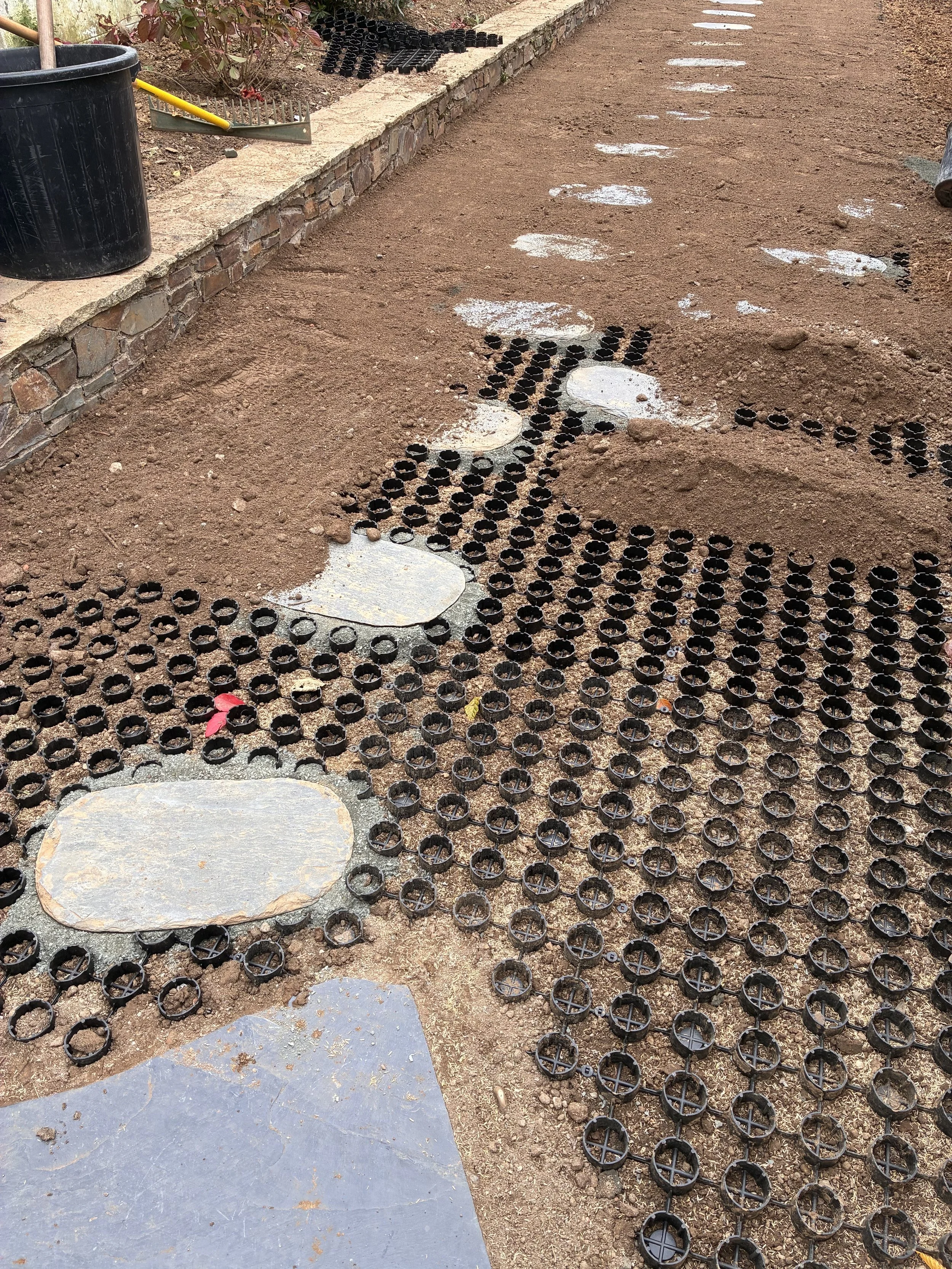 Installation en cours pour poser des dalles ou pavés sur un chemin, avec des rondelles noires en plastique pour la fixation, et quelques pierres plates déjà posées.