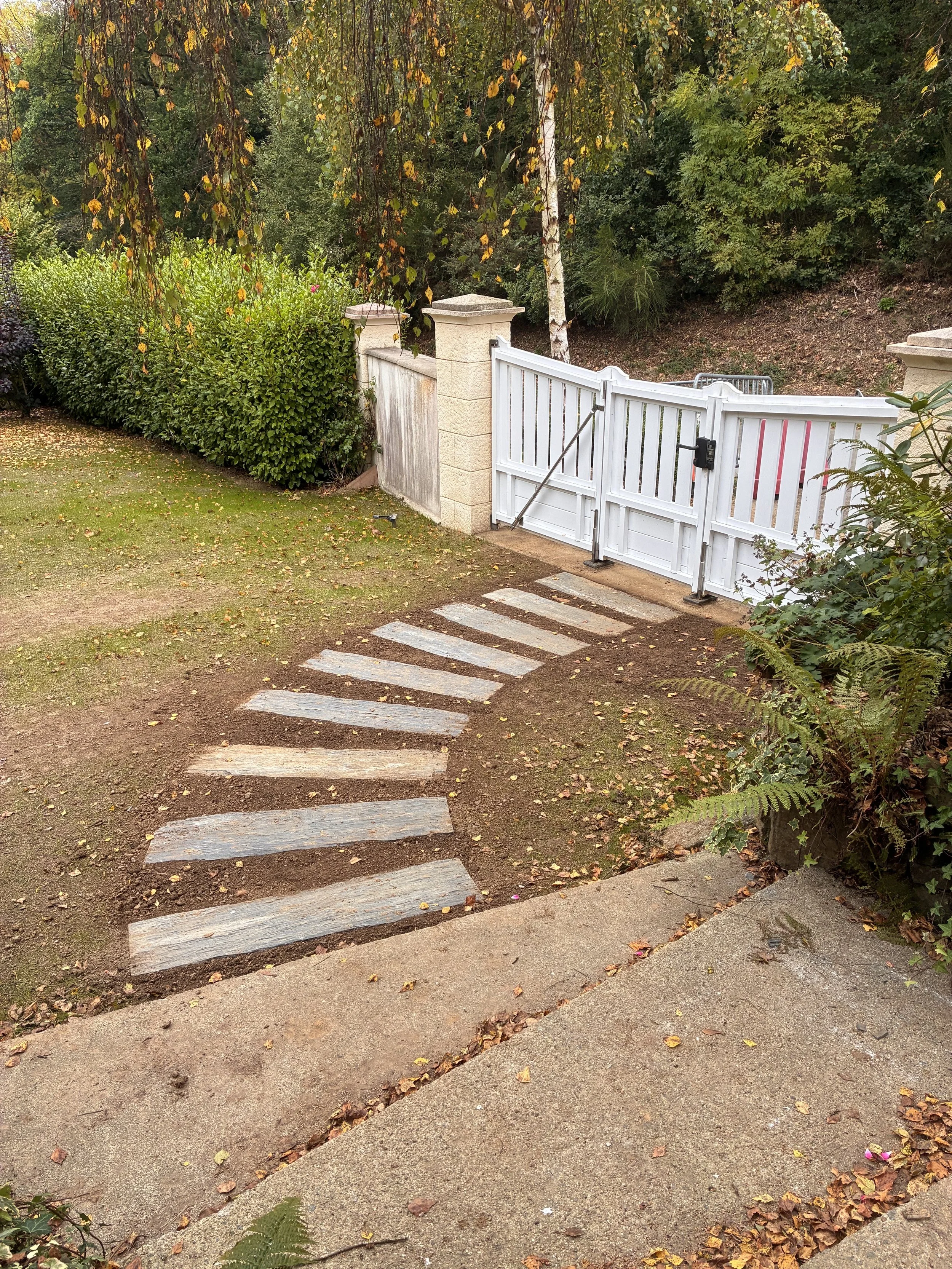 Chemin piétonnier en pierre menant à une porte blanche dans un jardin entouré d'arbres et de buissons, avec des feuilles tombées au sol.