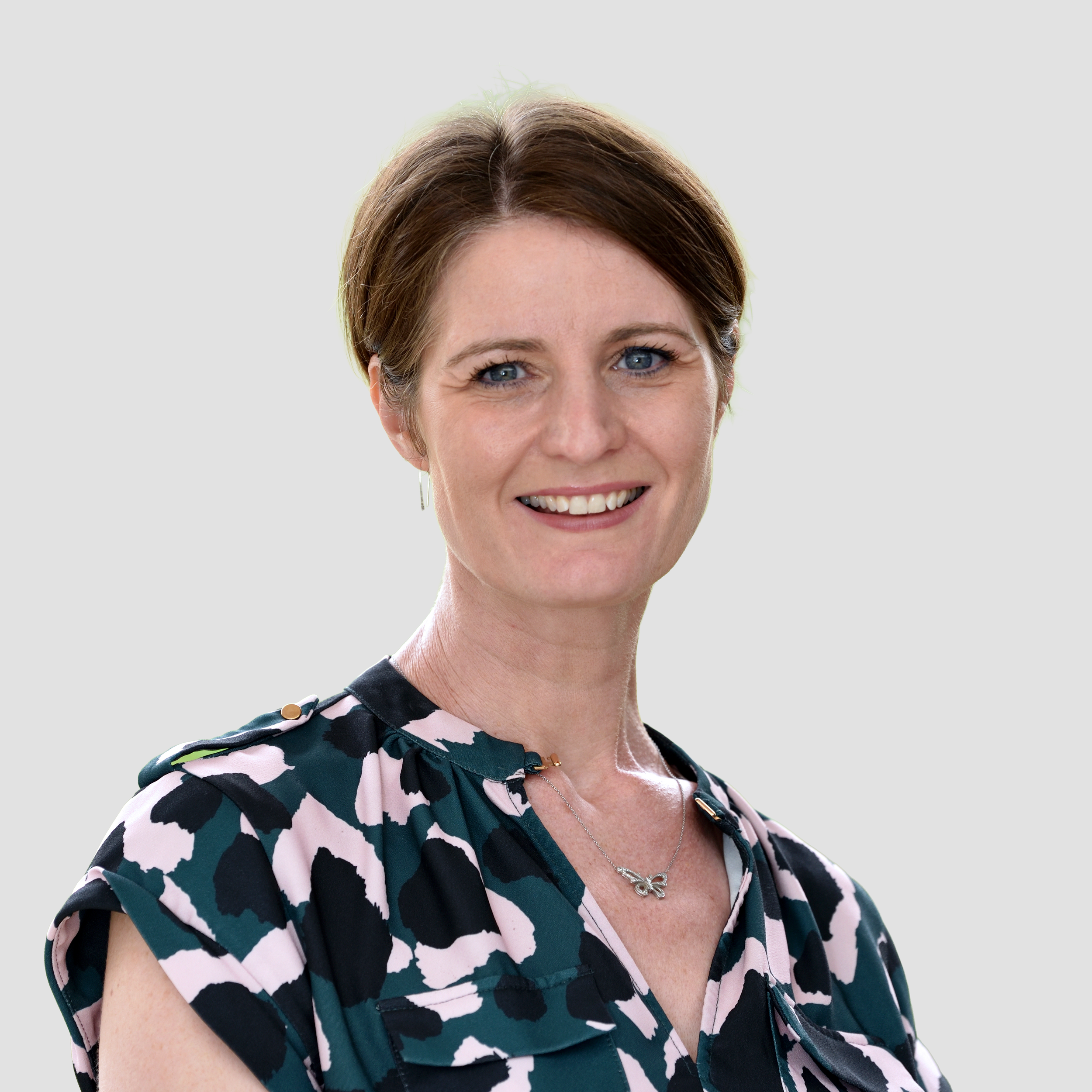 A smiling woman with short brown hair, blue eyes, and wearing a patterned blouse and a butterfly necklace.