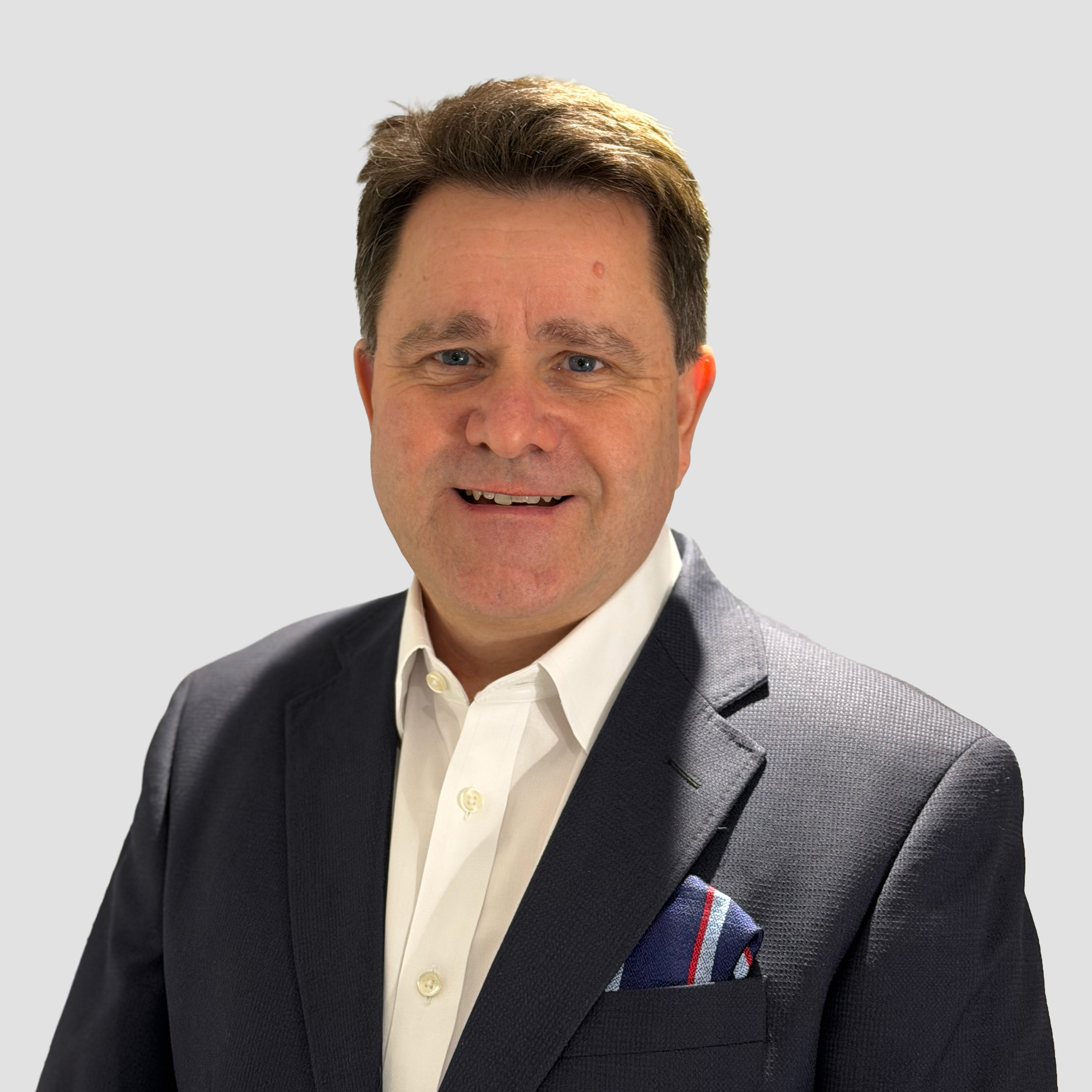 Headshot of a middle-aged man with short dark hair, wearing a dark suit jacket, white shirt, and a colorful pocket square, against a plain light gray background.