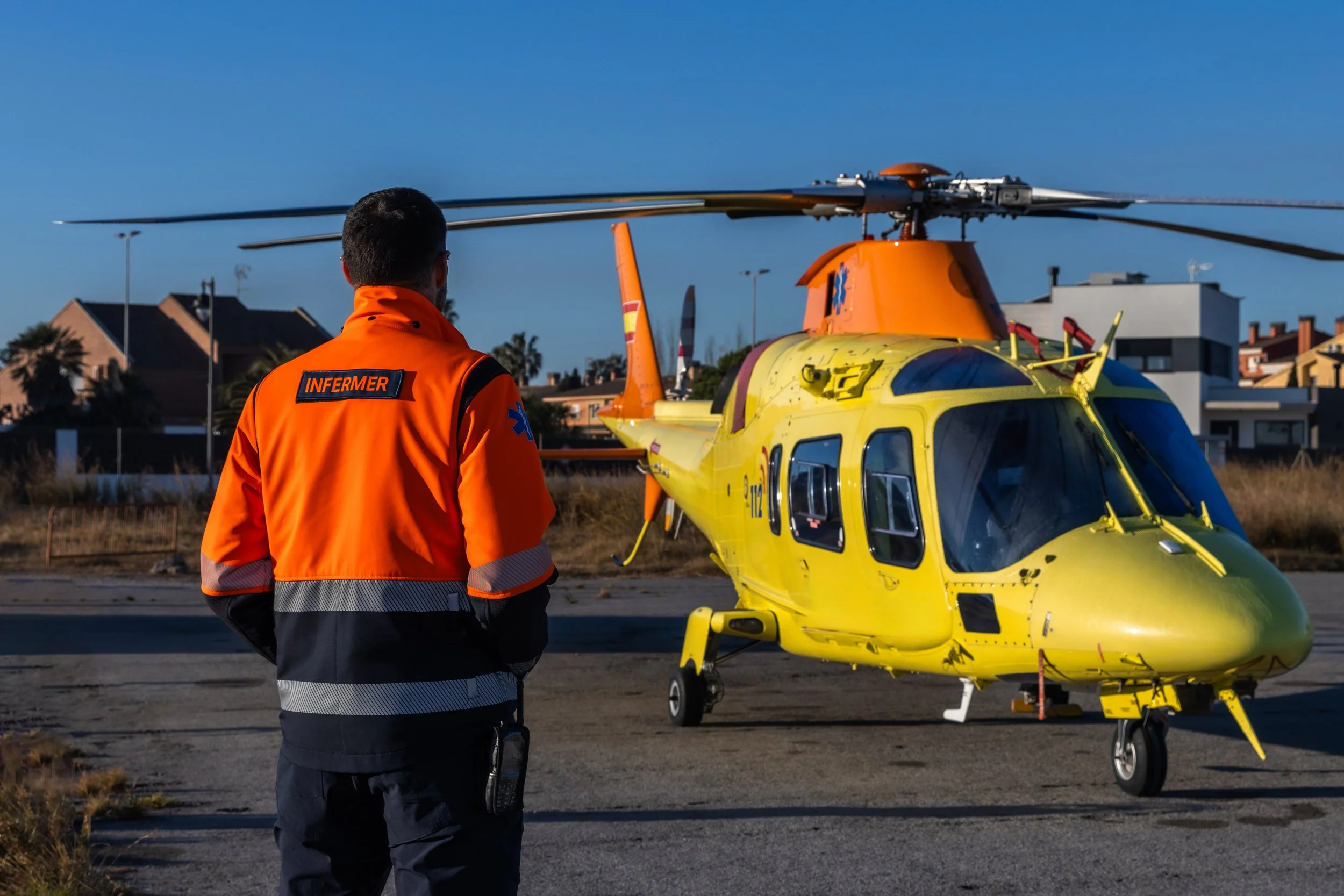 A person wearing an orange emergency medical technician (EMT) jacket standing near a yellow helicopter on a helipad, with residential buildings and a clear blue sky in the background.