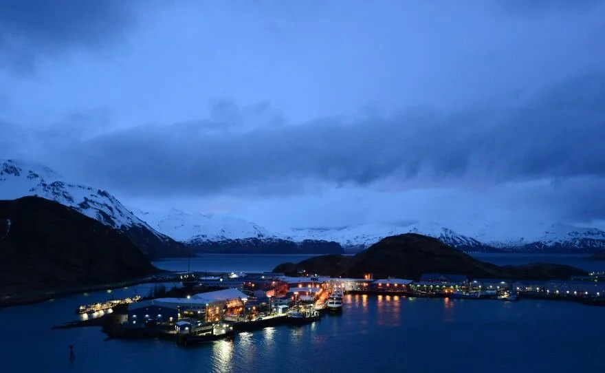 Night view of a harbor with boats, surrounded by snow-capped mountains and cloudy sky.