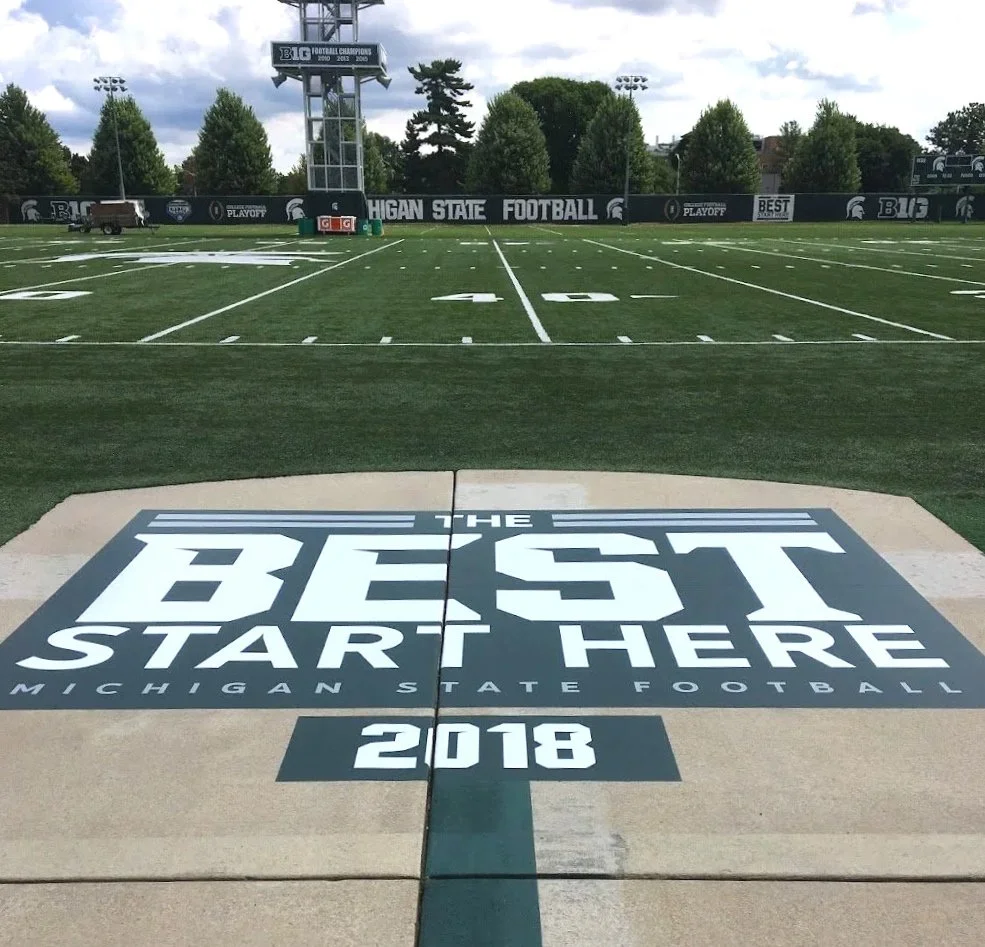 Michigan State Football field with a large graphic on the sidewalk that says "The Best Start Here 2018." The football field is empty, with trees and a cloudy sky in the background.