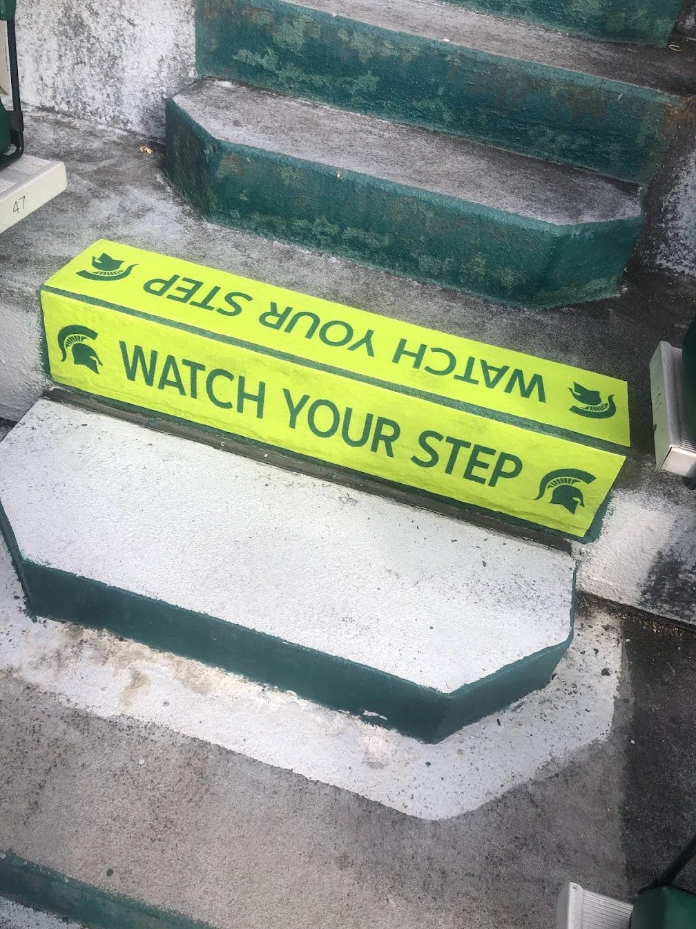 Bright yellow warning sign on the ground that says 'WATCH YOUR STEP' with a Spartan helmet logo, placed at the bottom of a green painted outdoor concrete stairs.