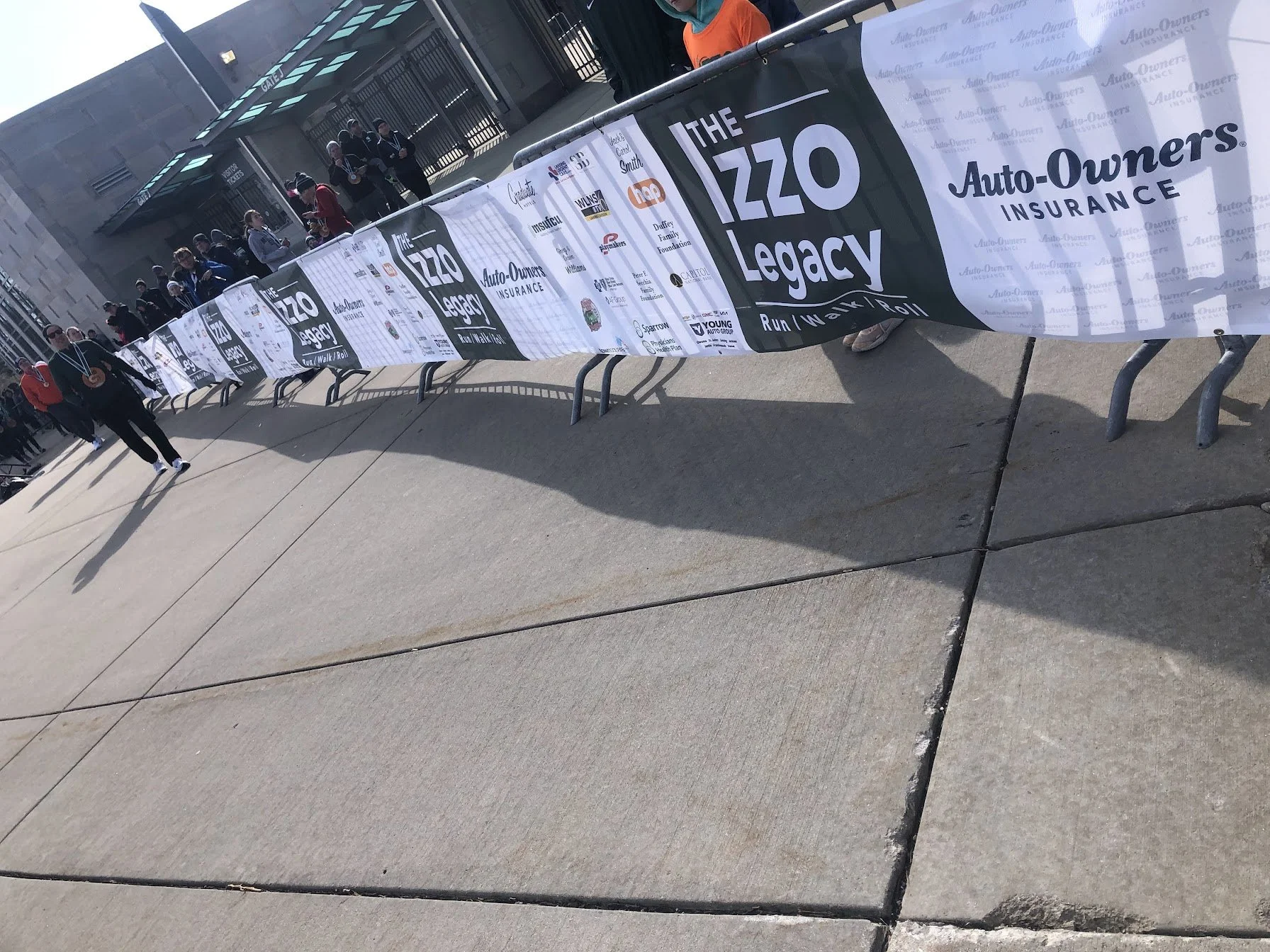 A group of people standing behind a barrier decorated with banners for the 'IZZO Legacy Run/Walk/Roll' event, outside a building with a glass entrance and a metal roof structure, on a sunny day.