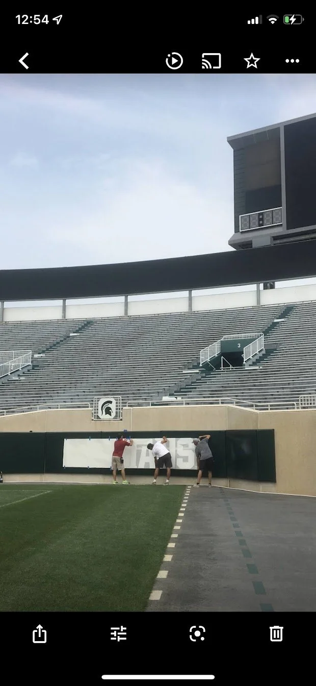 Three people installing or fixing a large sign on a wall inside a stadium.
