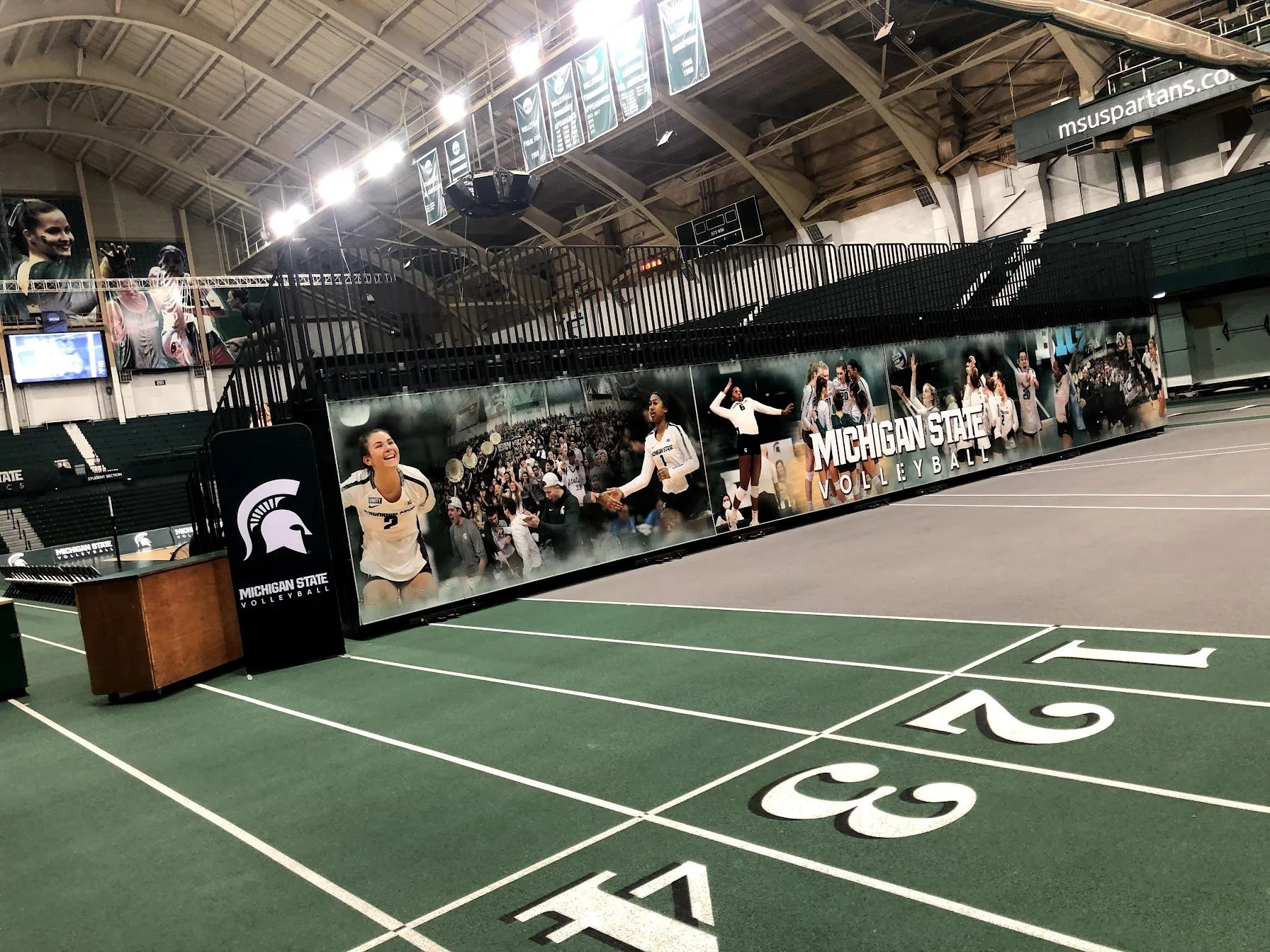 An indoor volleyball court with green flooring and white markings. A Michigan State Volleyball banner with images of players and fans decorates the enclosed area. The arena is well-lit, with banners hanging from the ceiling, and empty seating surrounds the court.