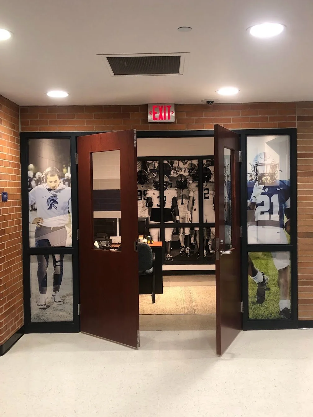 Entrance to a sports office with large football player posters on glass doors, showing players in football uniforms and helmets, along with a display of football jerseys and helmets inside.