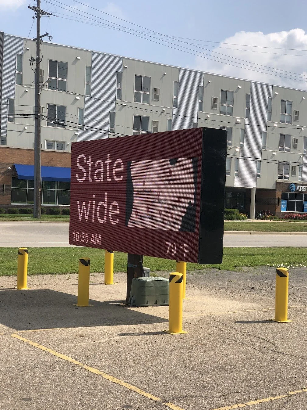 An electronic billboard displays the words "State wide" and the time as 10:35 AM, with the temperature showing 79°F. In the background, there is a modern apartment building with a white facade, and a small AT&T store with a blue awning. Yellow bollards are placed around the billboard for protection.