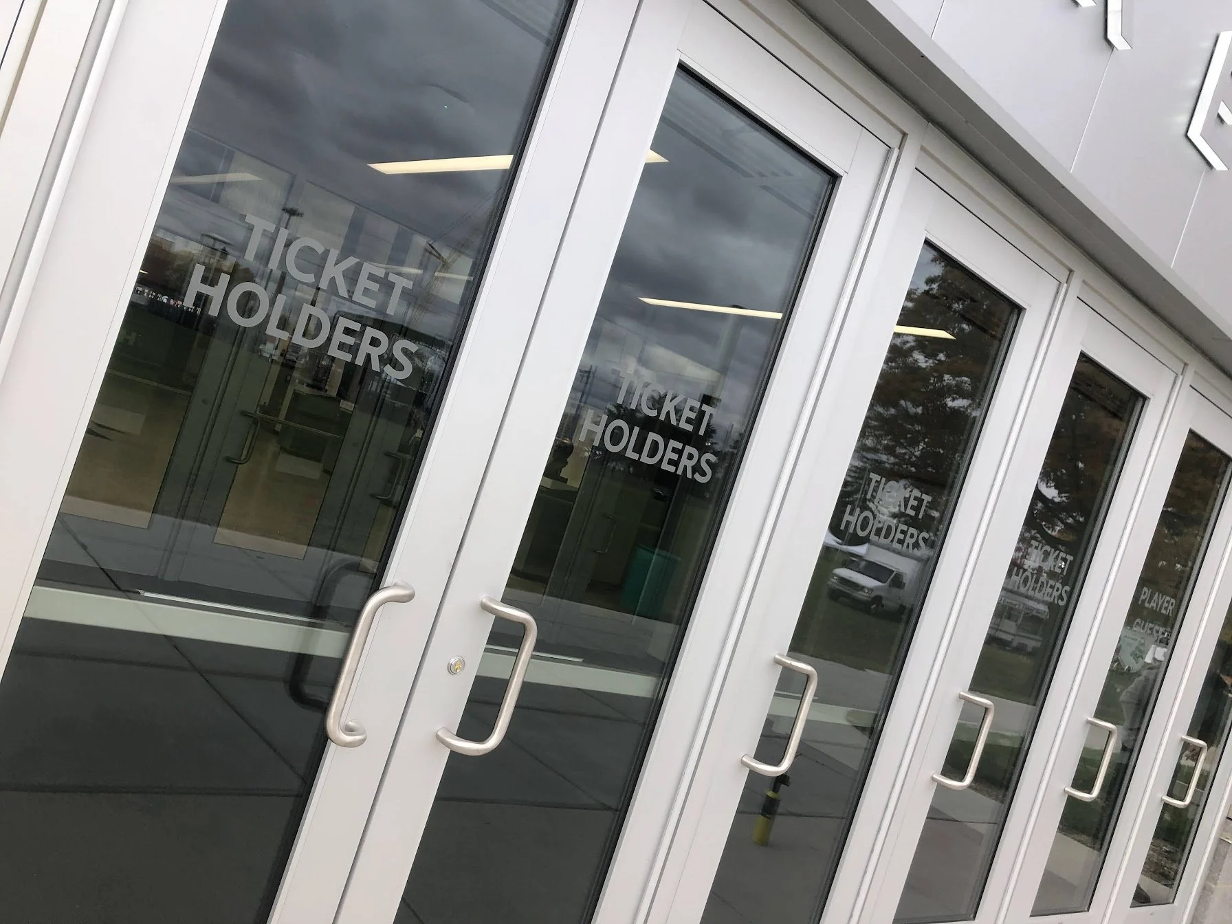 Glass double doors with metal handles leading into a building, with 'TICKET HOLDERS' written on the glass, and reflections of trees, cars, and a cloudy sky.