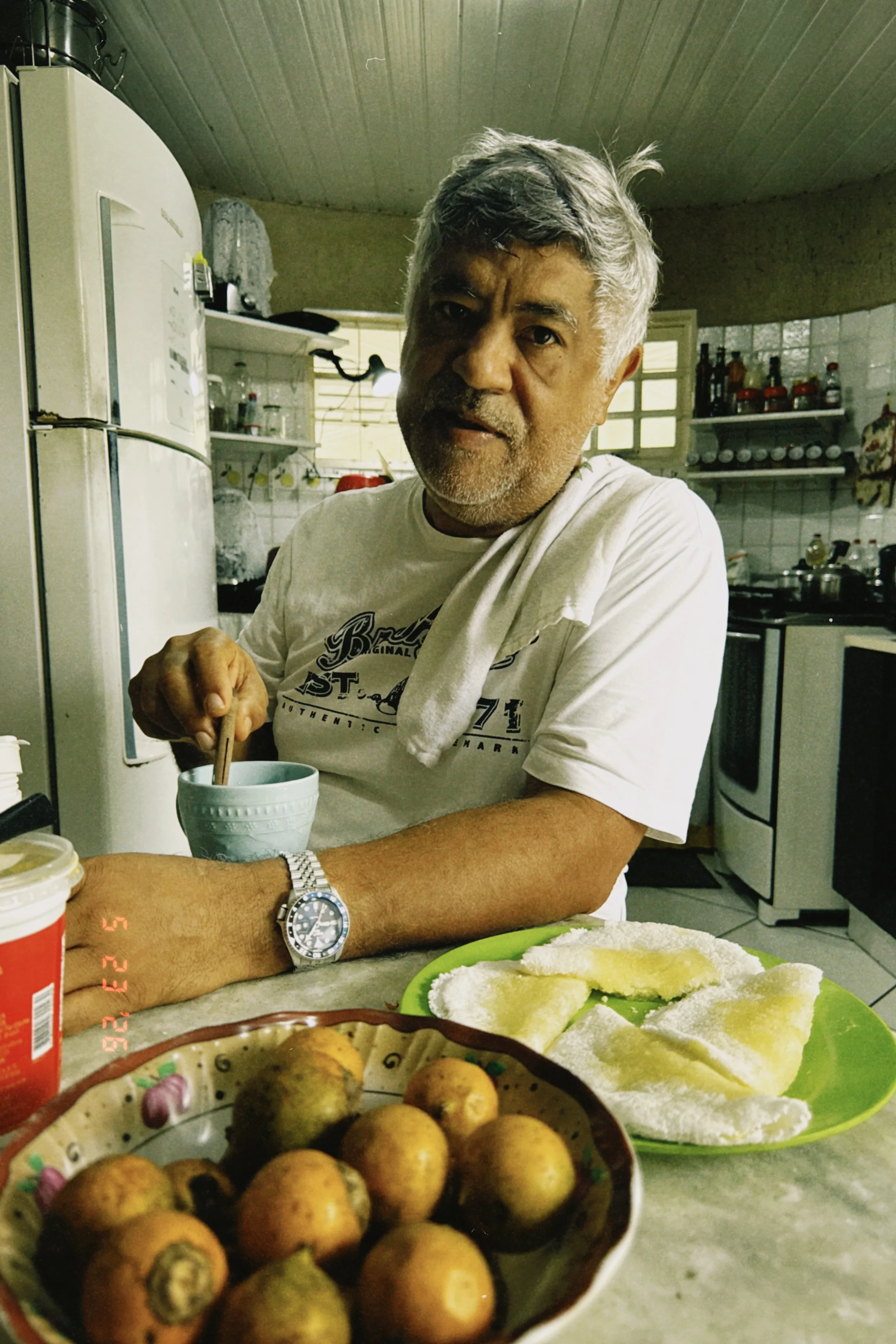 A middle-aged man with gray hair and a beard sitting at a kitchen table, stirring a cup with a spoon. The kitchen has a white refrigerator, shelves with jars, and a stove in the background. On the table, there is a green plate with slices of fruit or