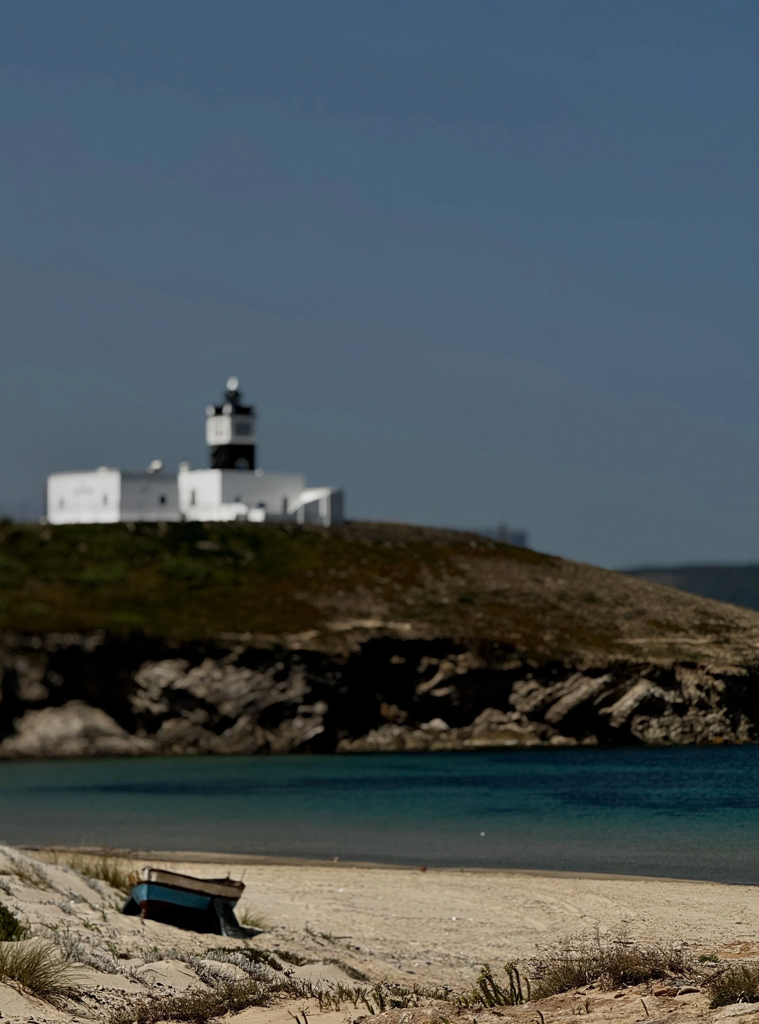 A small boat rests on a sandy beach with grass patches, with a large body of water beyond, and a distant hillside with a white lighthouse atop it.