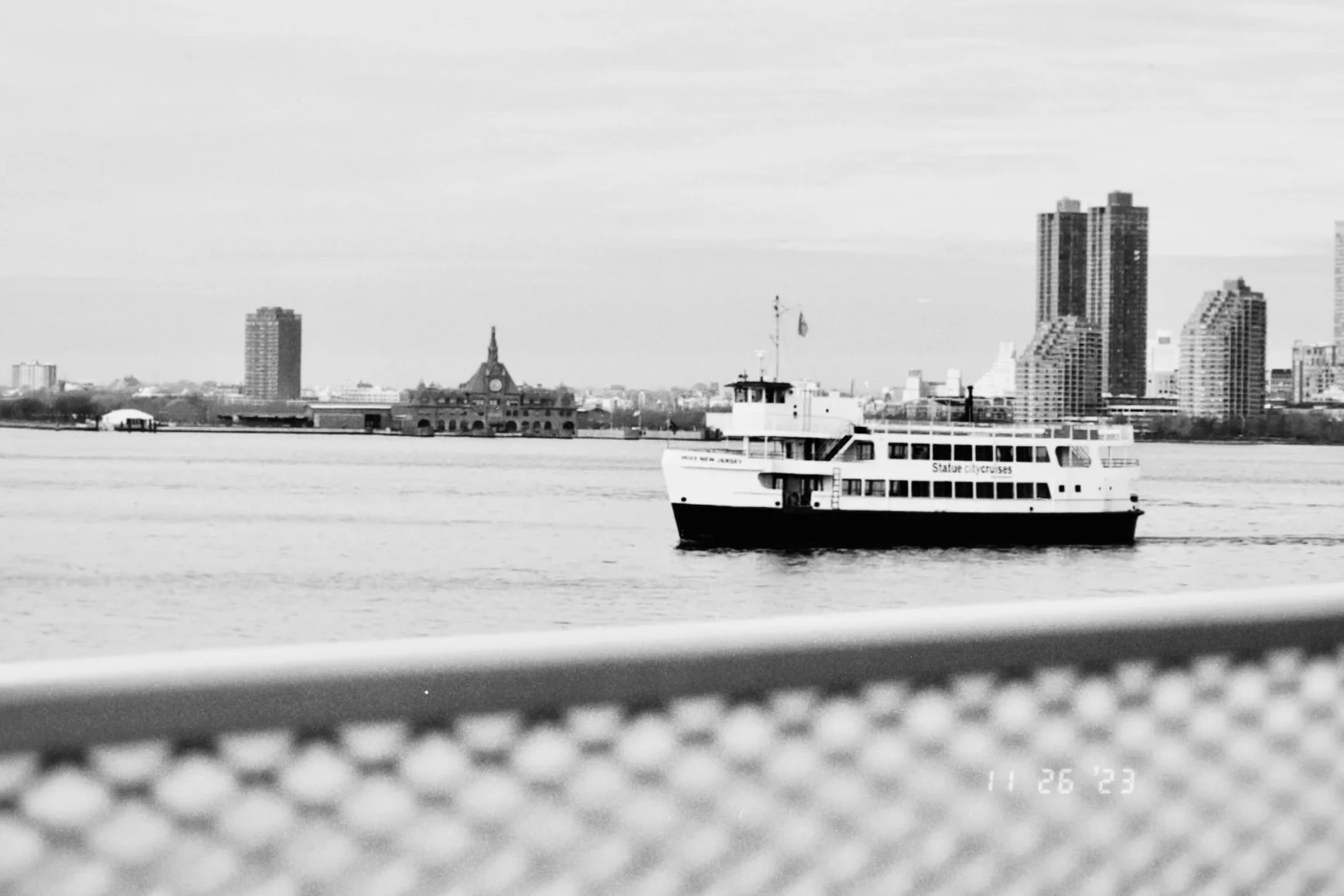 Black and white photo of a boat on a river with city buildings in the background, including a clock tower and high-rise apartments.